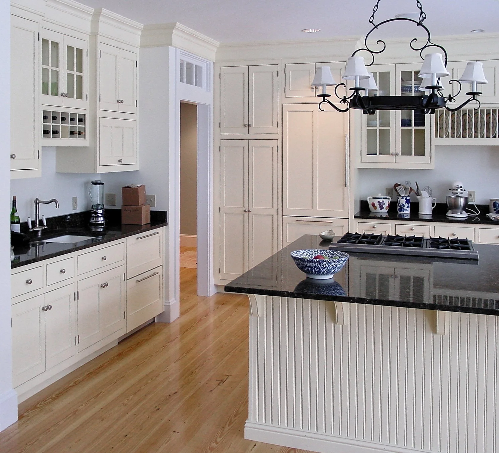 A modern kitchen with white cabinets, black countertops, and hardwood flooring. An island with a dark countertop is in the center, and a chandelier hangs above. Some dishes and small kitchen appliances are on the counter.