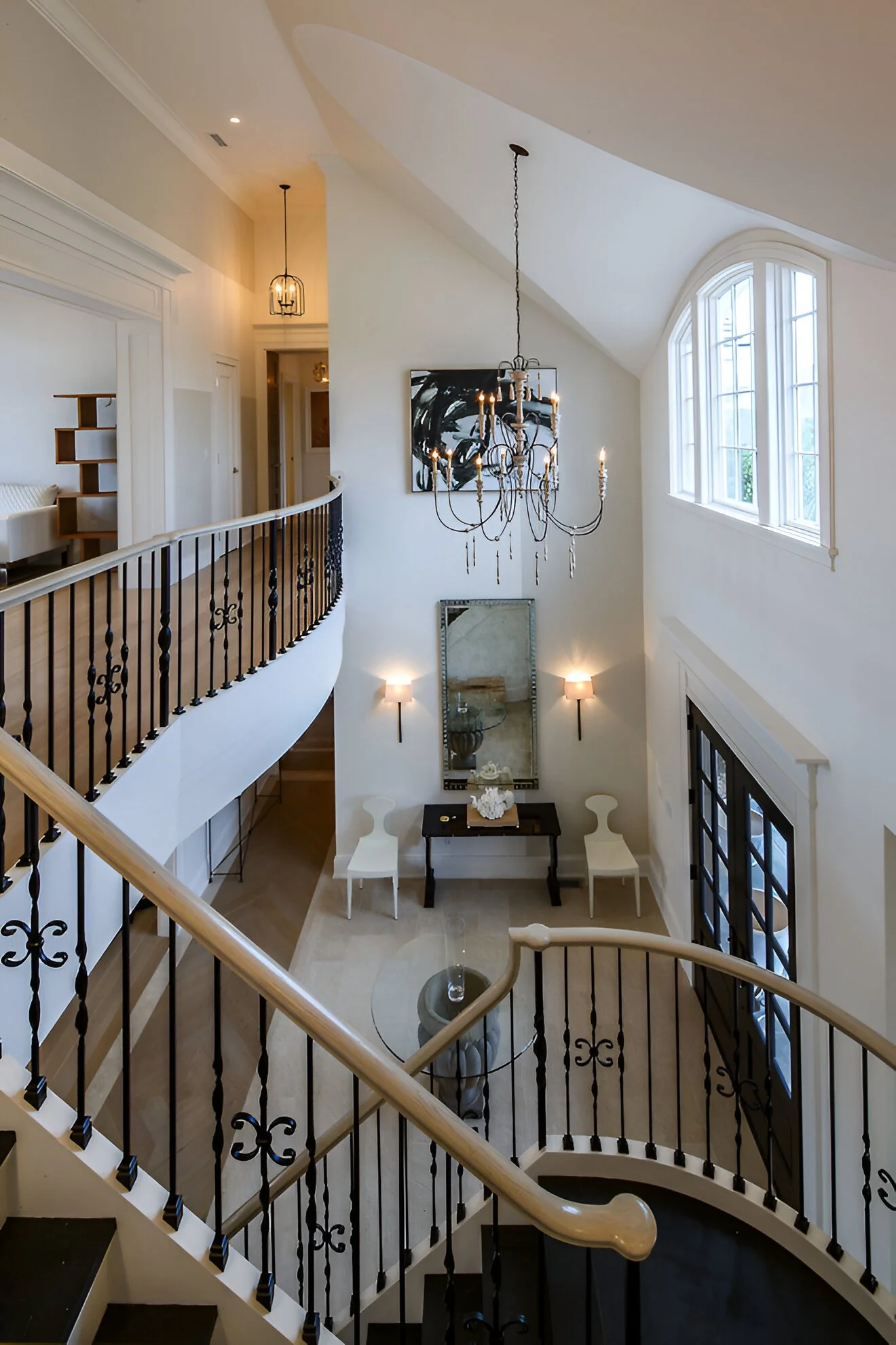 Interior view of a modern, elegant staircase area with a black wrought iron railing, a chandelier hanging from the ceiling, large arched windows, and wall sconces illuminating the space.