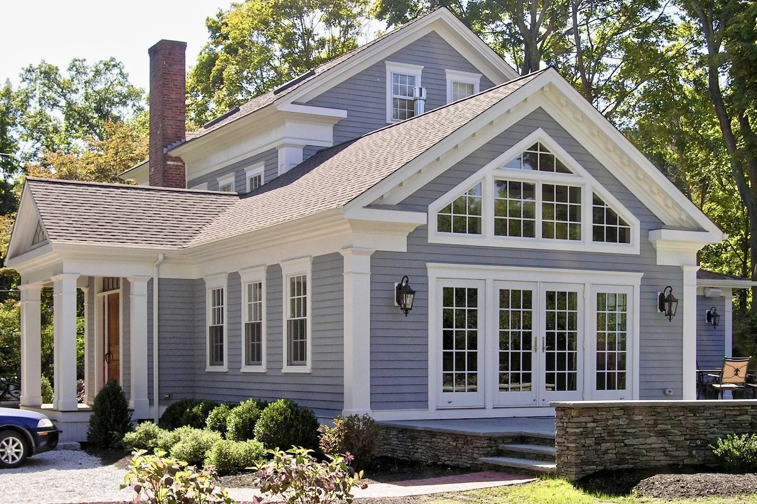A gray two-story house with white trim, large front windows, and a stone foundation, surrounded by trees and greenery.