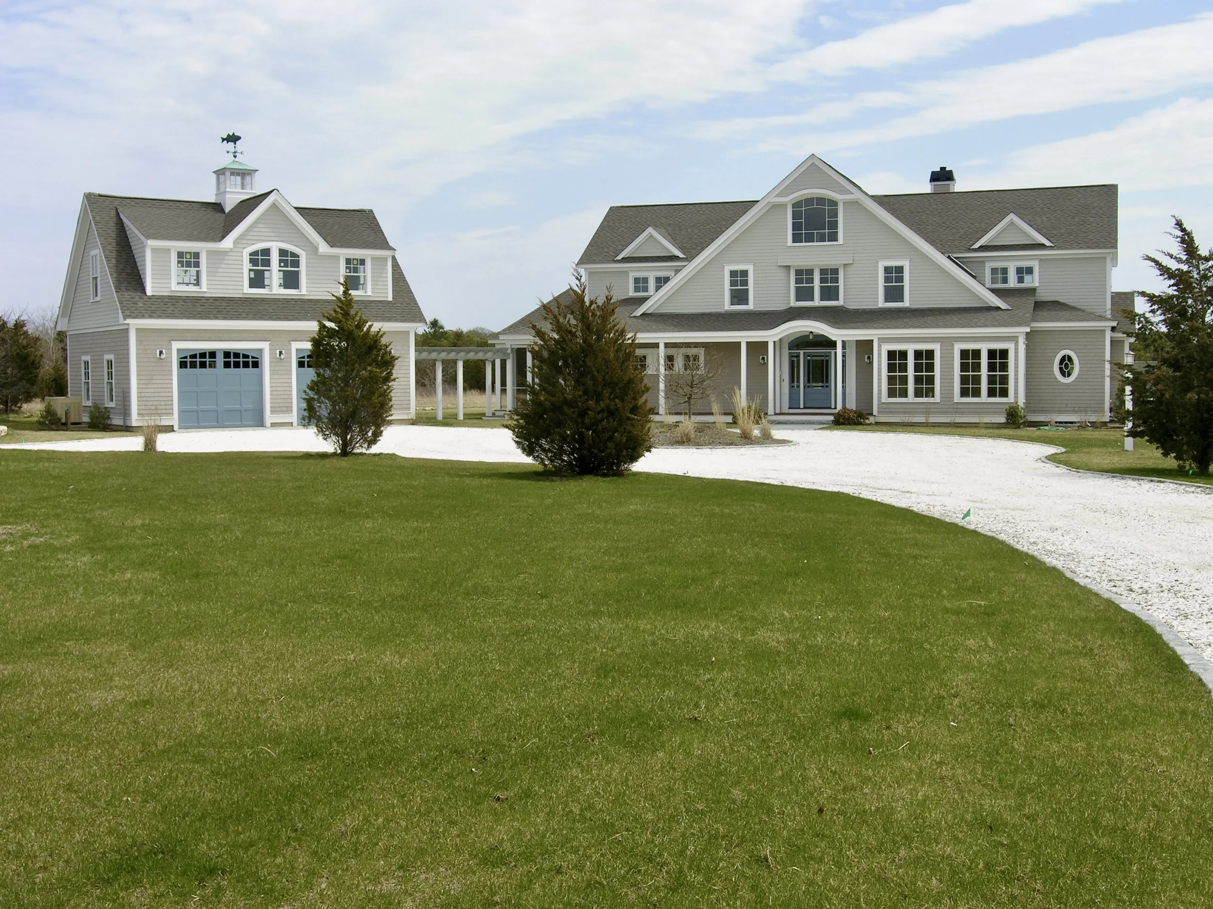 Large gray house with blue door and attached garage, surrounded by a well-manicured lawn and trees on a cloudy day.
