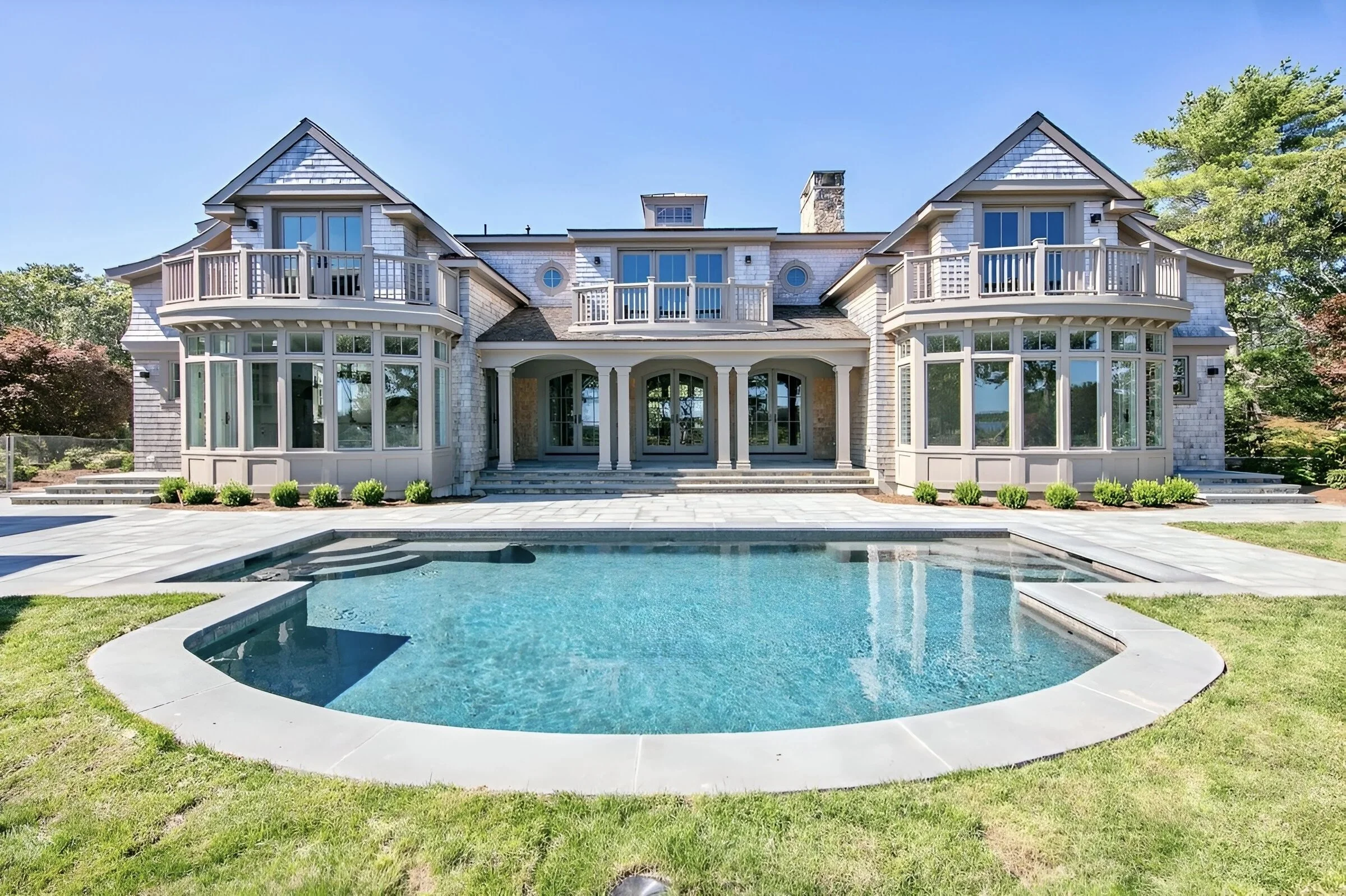 Luxury home with multiple levels, balconies, and large windows, situated behind a curved gunite swimming pool and green lawn, under a clear blue sky.