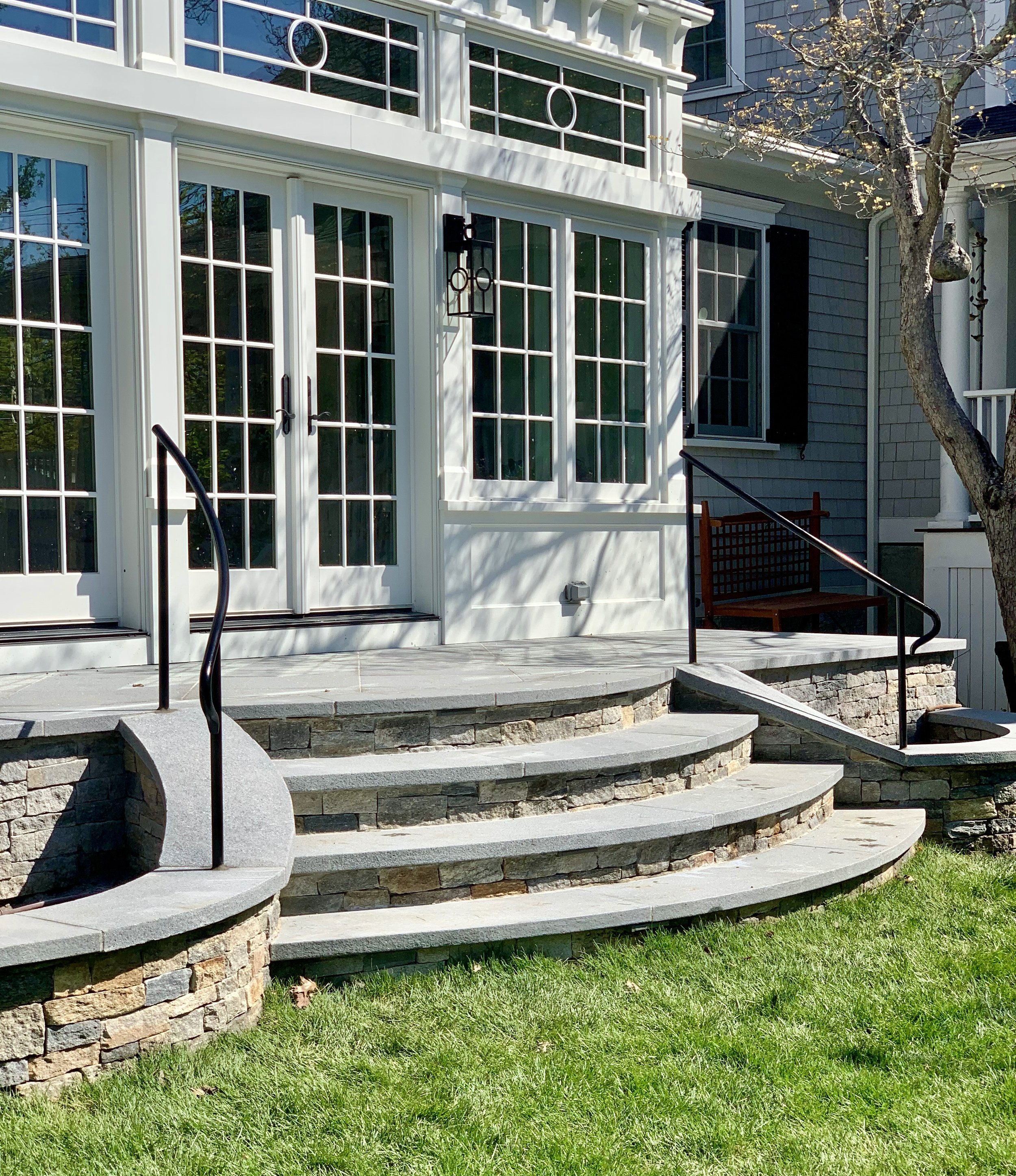 Exterior view of a sunroom with a curved stone staircase leading up to a porch with white French doors and large windows. There are black metal railings and a wooden bench on the porch. A tree with budding leaves is visible on the right side.