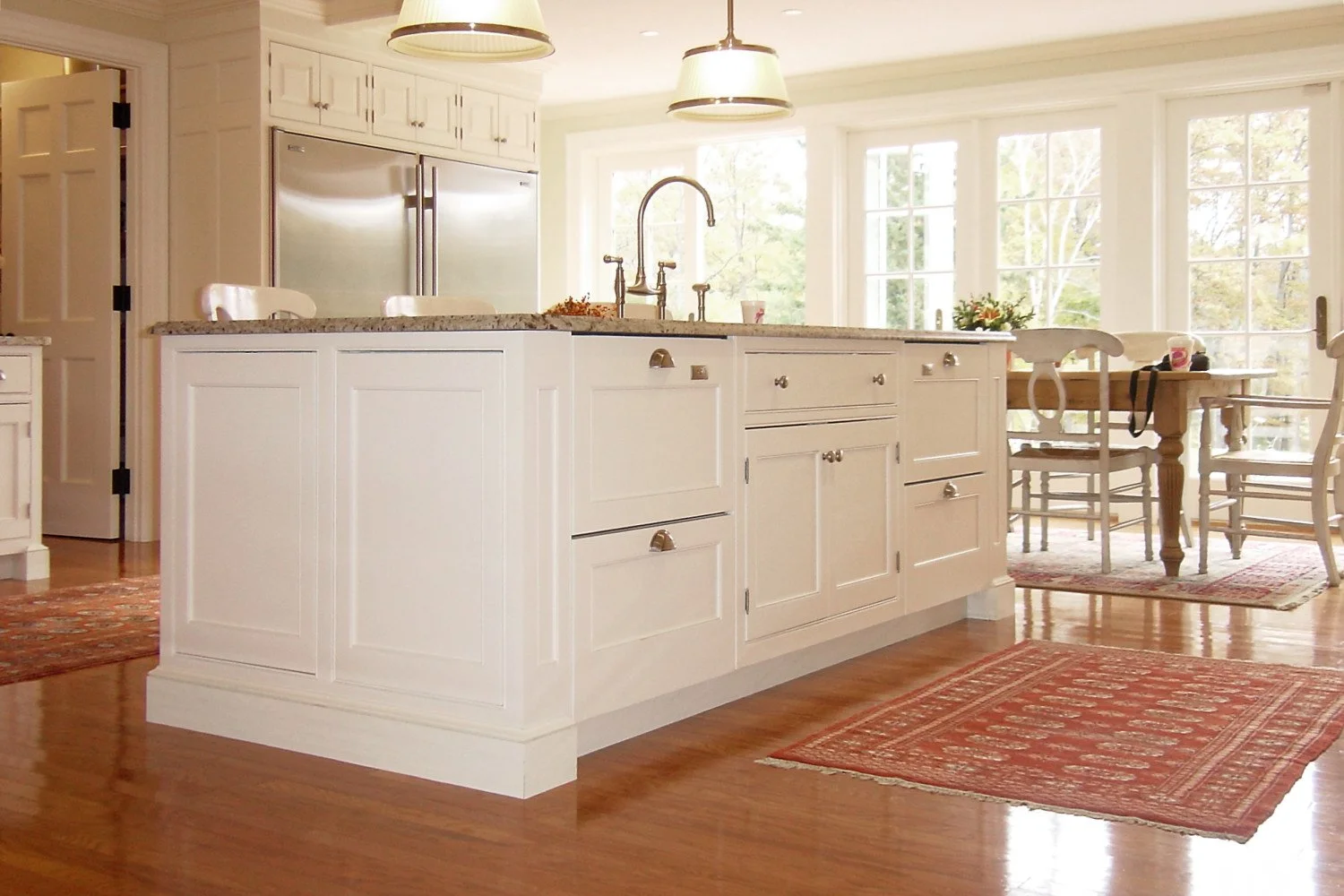 A bright kitchen with white cabinetry, a large island with a granite countertop, and a sink with a gooseneck faucet. There are hanging pendant lights above the island, and large windows in the background letting in natural light. A wooden dining tabl