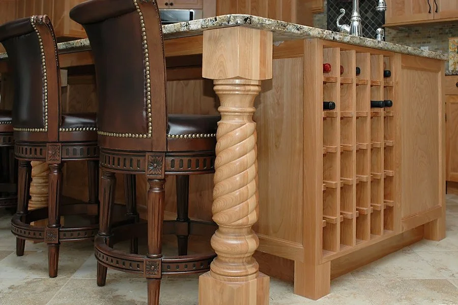 A kitchen island with a granite countertop, supported by a decorative twisted wood leg. Next to the island are two barstools with dark wood legs and leather upholstery. The island has a built-in wine rack with several bottles stored horizontally.