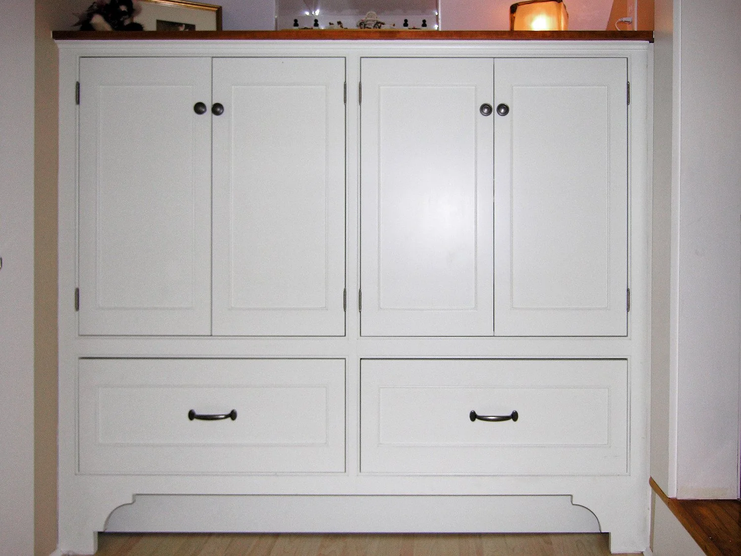 White wooden cabinet with two upper doors and two lower drawers, black handles, located in a room with beige walls and hardwood floor.