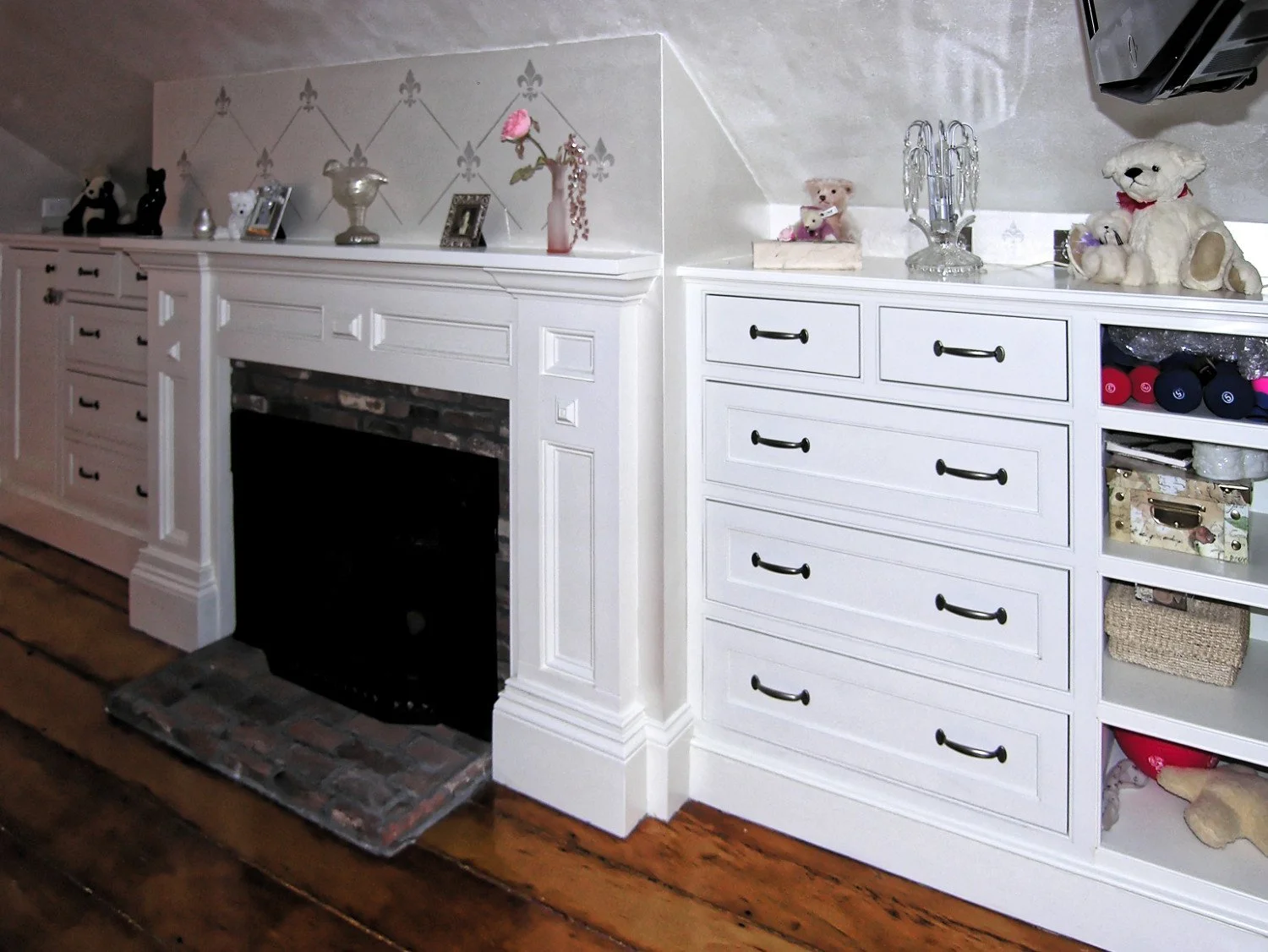 White wooden cabinetry with drawers and shelves, a brick fireplace with a mantel decorated with various plush toys, framed photos, and decorative items, under a wall with a diamond pattern wallpaper.