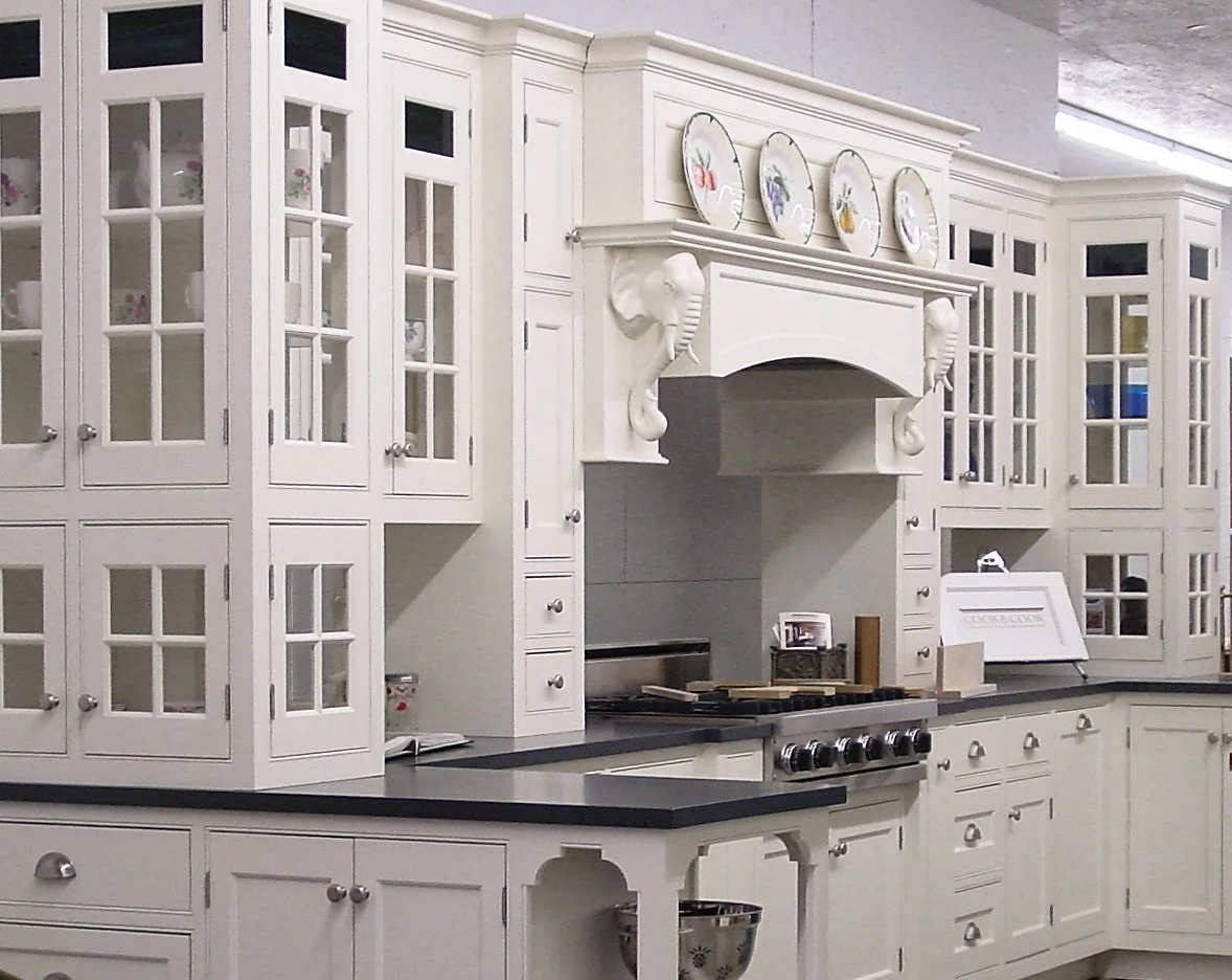 A white kitchen with glass-front cabinets, a stove under a decorative hood with elephant motifs, and a black countertop. Decorative plates with floral designs are displayed above the hood.