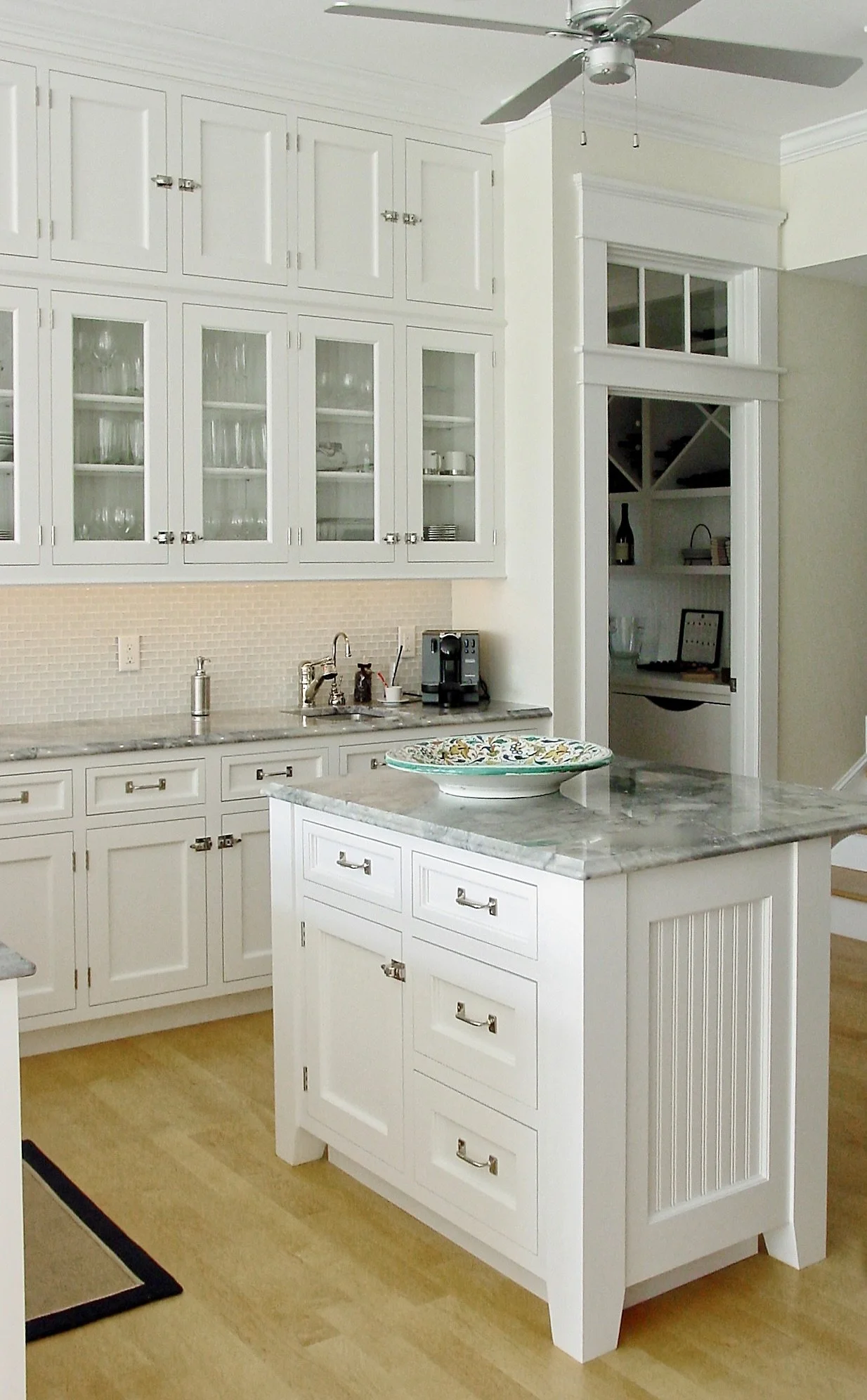 White kitchen with marble countertops, glass-front cabinets filled with glassware, a coffee machine, and a decorative plate on a kitchen island.