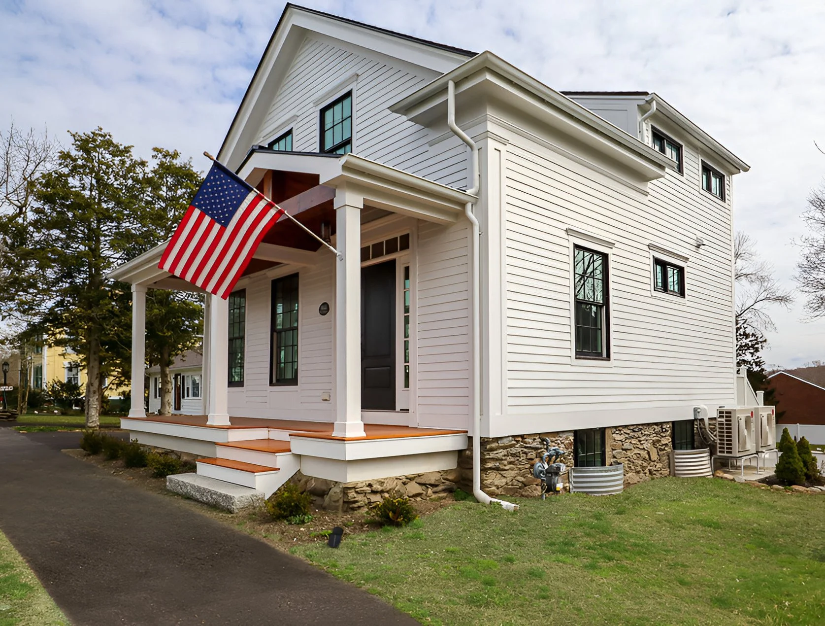 A white two-story house with black window frames, a small porch with white columns, and an American flag hanging from a pole near the porch. The house has a stone foundation and green lawn with small shrubs, with other houses and trees visible in the background.