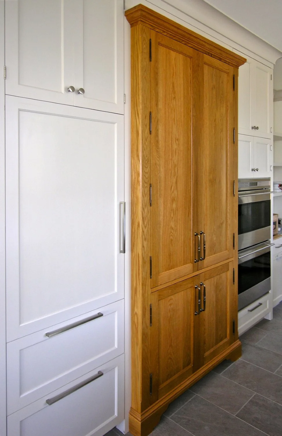 A wooden oak pantry cabinet with double doors standing between white cabinets and kitchen appliances in a kitchen.