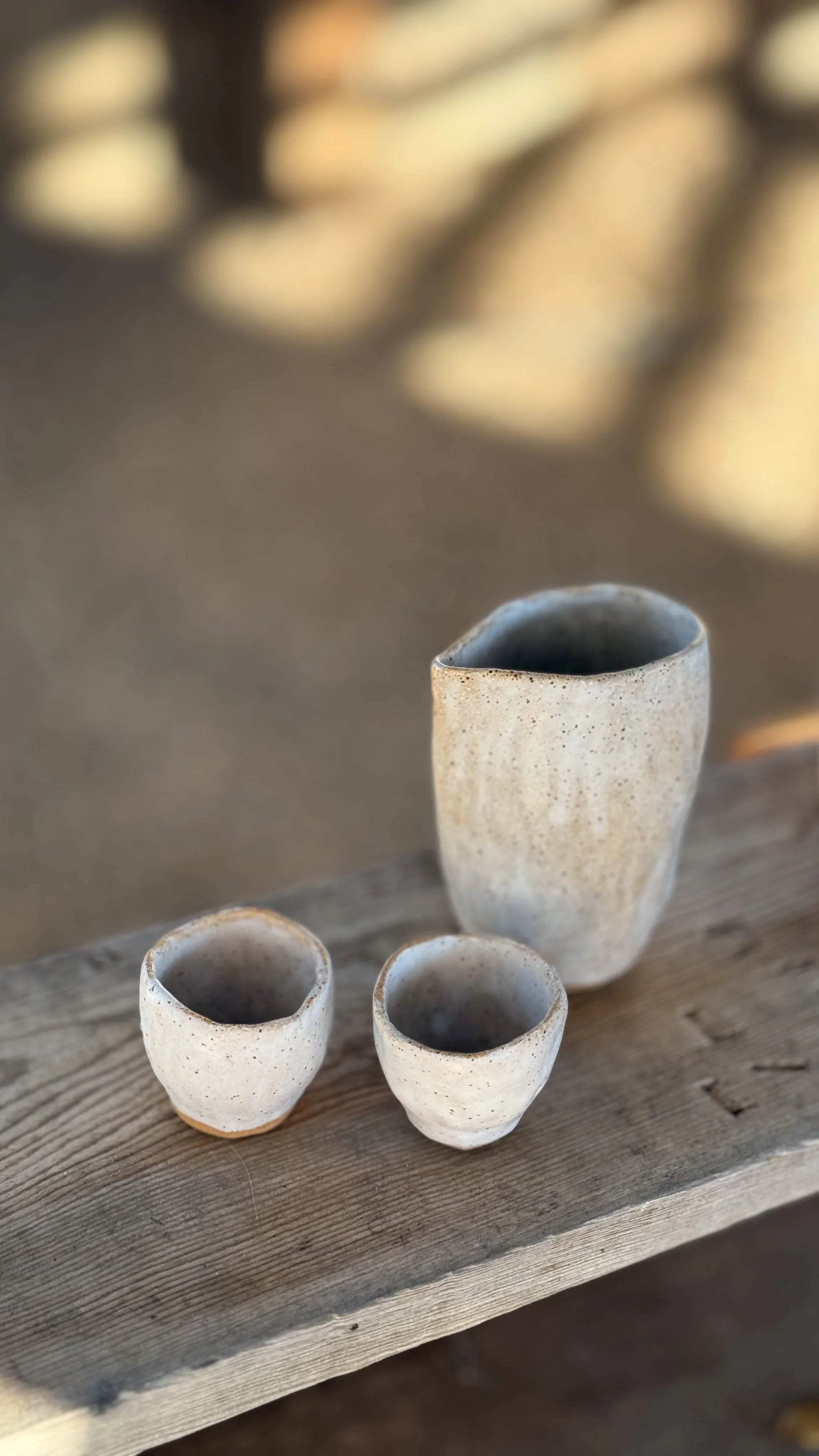 Three small pottery cups and one larger pottery cup on a wooden plank, outdoors.