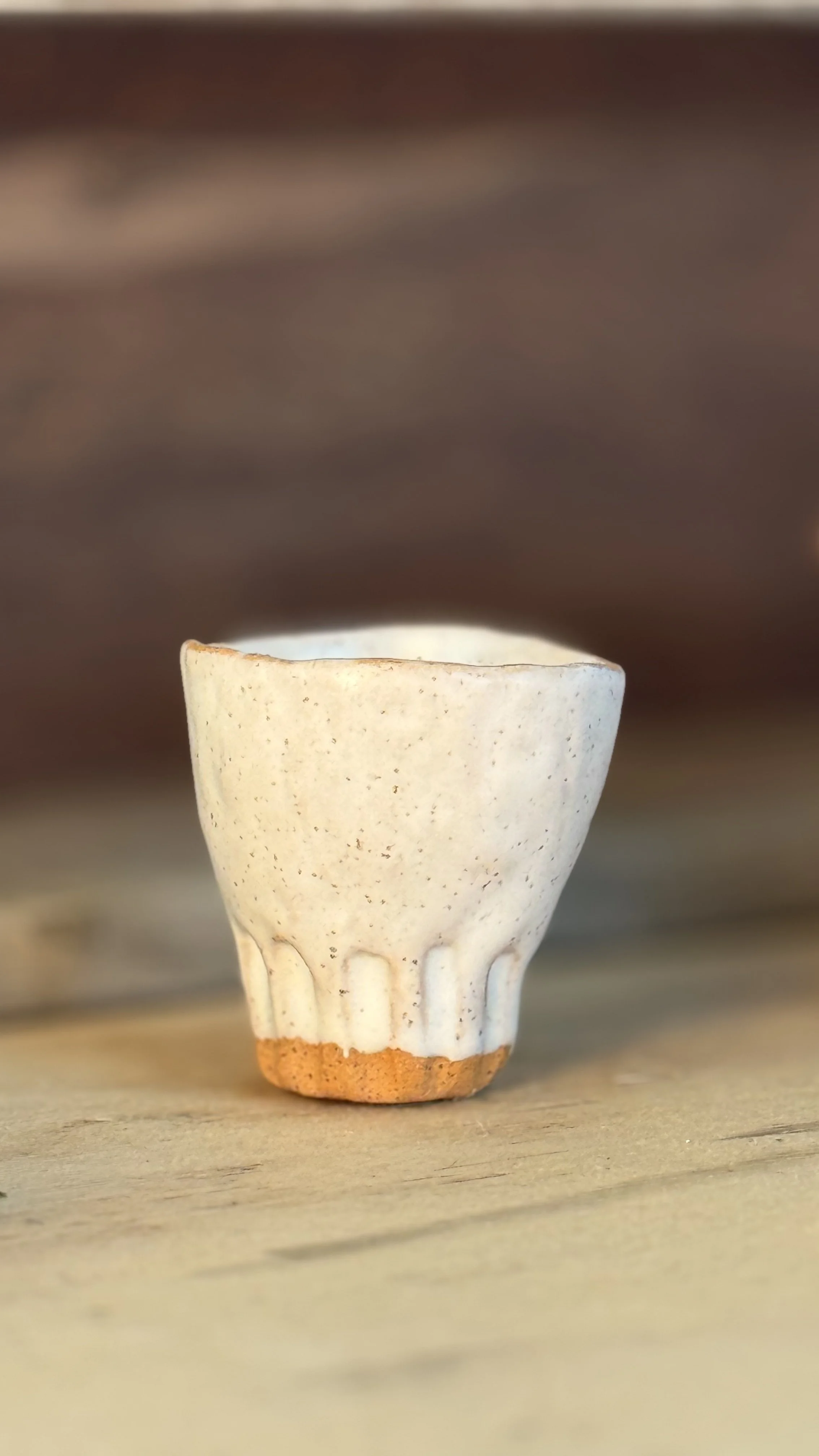 Close-up of a small, cream-colored ceramic cup with vertical grooves at the base, placed on a wooden surface with a blurred dark background.