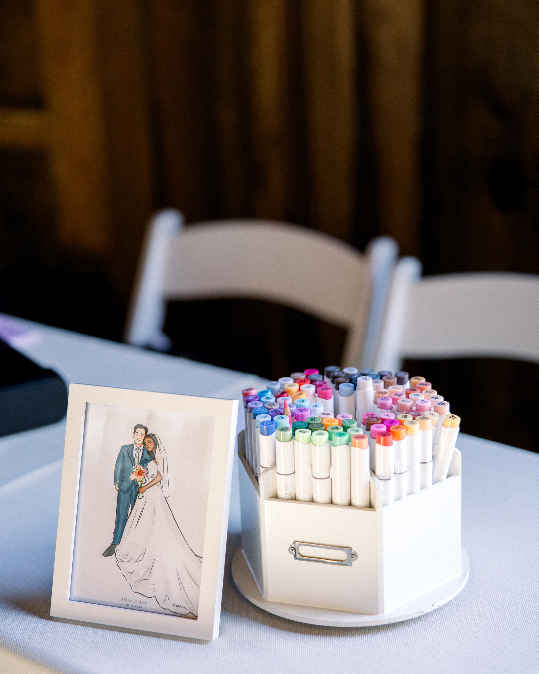 A framed illustration of a bride and groom on a table next to a container of multicolored markers at a wedding reception.
