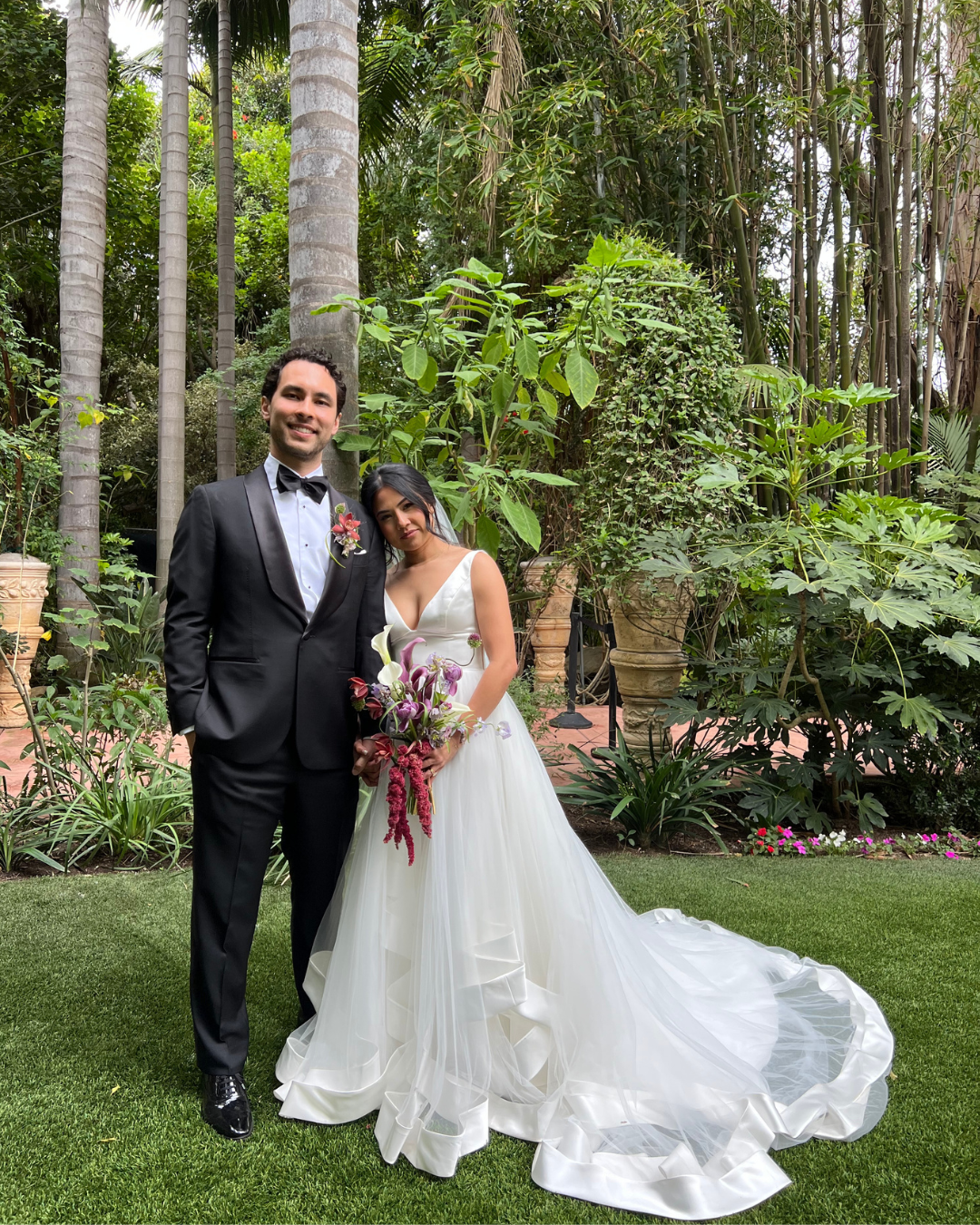 A bride and groom standing together outdoors in a lush garden, with the bride holding a bouquet of flowers and both dressed in wedding attire.