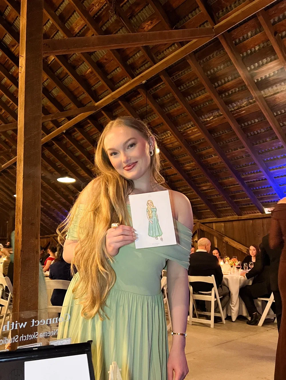 A young woman with long, wavy red hair wearing a light green off-shoulder dress is standing in a rustic barn-style venue. She is smiling and holding a drawing of a woman in a green dress. There are tables with people in the background, and the ceilin