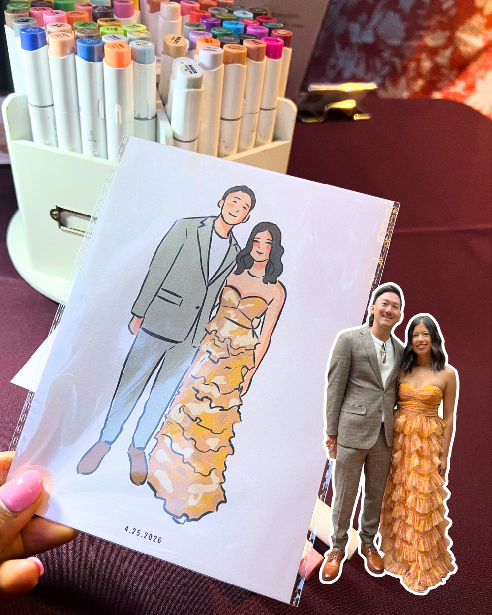 A hand holding a personalized illustration of a man and woman in formal attire, alongside a printed photo of the same couple. In the background, there's a container of colorful markers on a burgundy tablecloth, and a blurred red and orange backdrop.