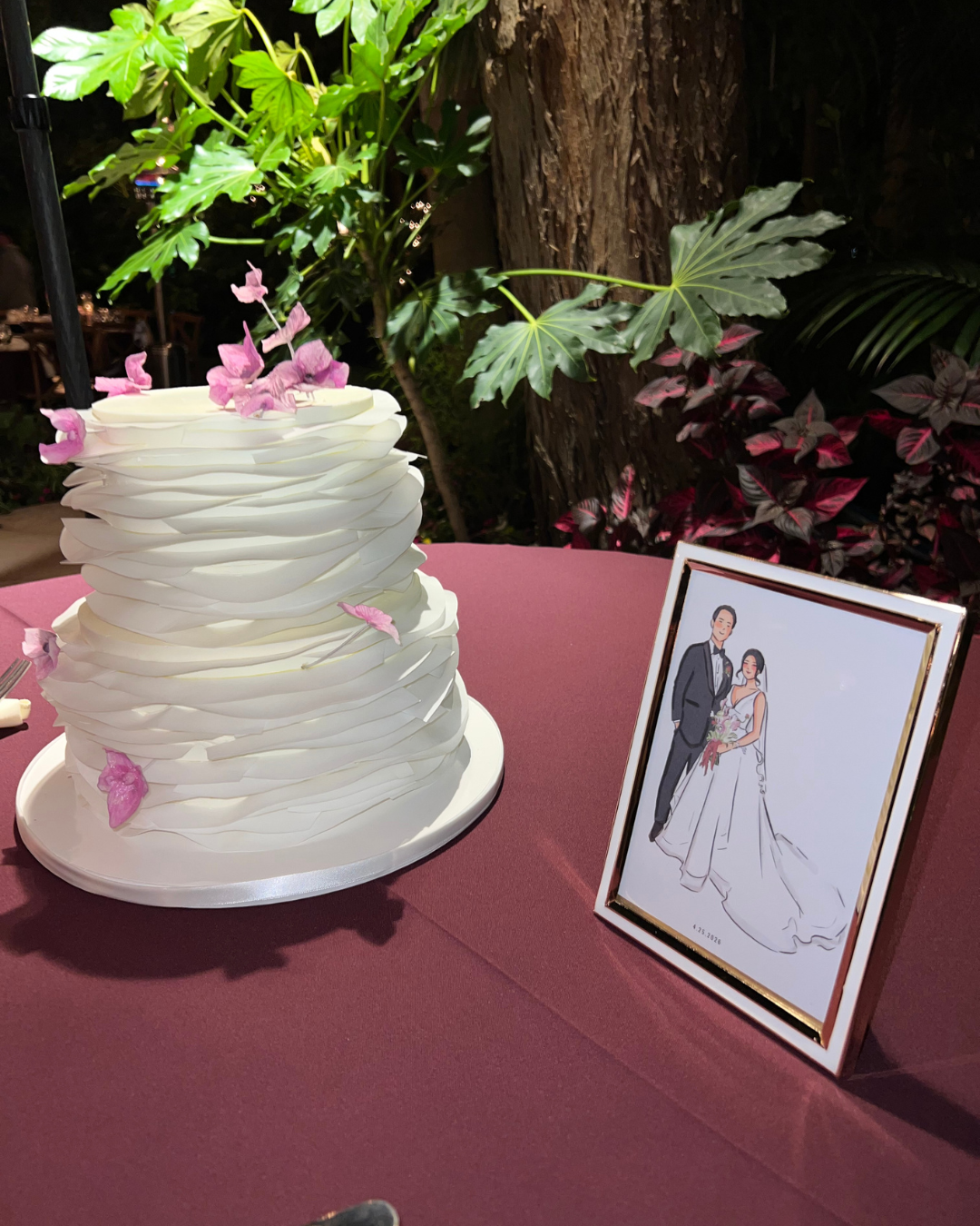 A layered white wedding cake with pink flowers on top, set on a pink tablecloth, with a framed illustration of a bride and groom next to it, surrounded by greenery.