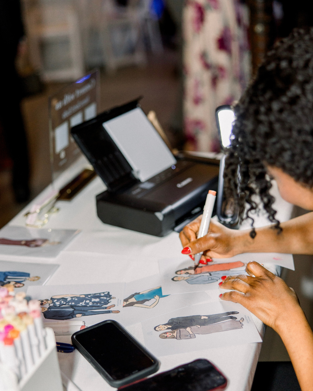 A person with curly hair drawing fashion illustrations on paper at a table with a printer, smartphone, and supplies.