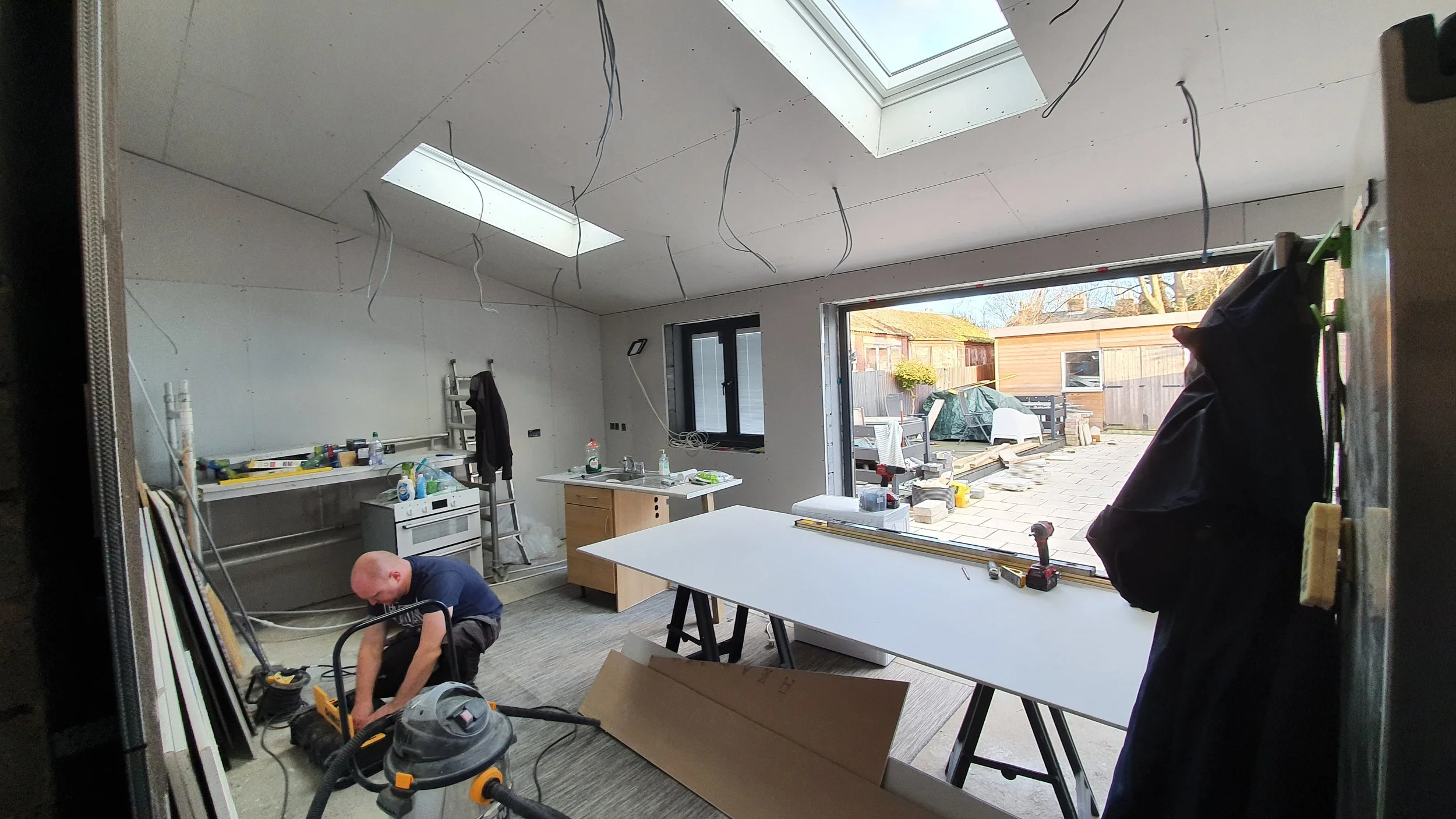 Interior of a house under construction or renovation with exposed ceiling wires, skylights, and a large sliding door leading to an outdoor patio. Construction workers are installing fixtures and working on the space.