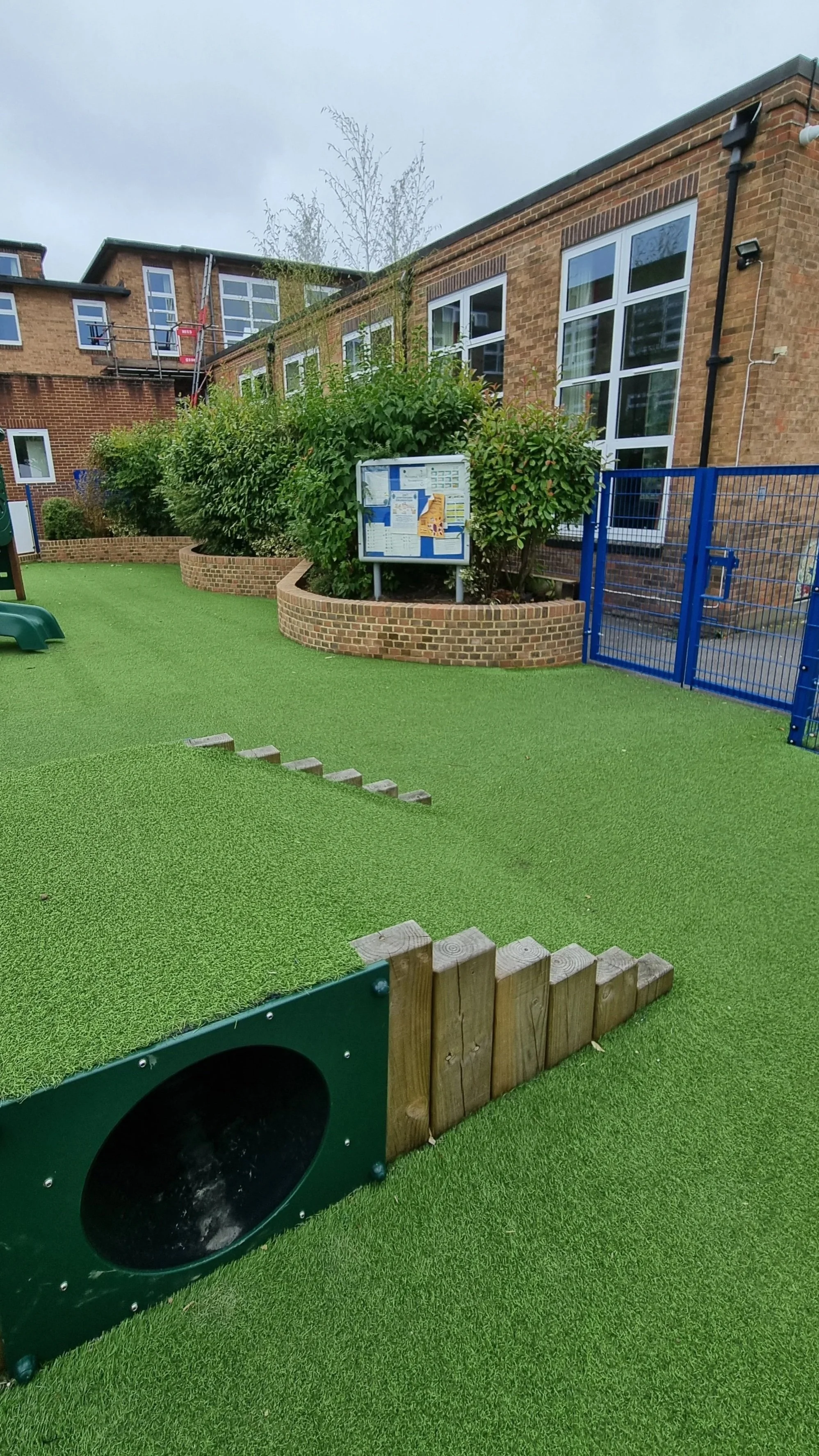 Playground area with artificial grass, wooden border, green tunnel, bushes, brick building with windows, blue gate, and notice board.