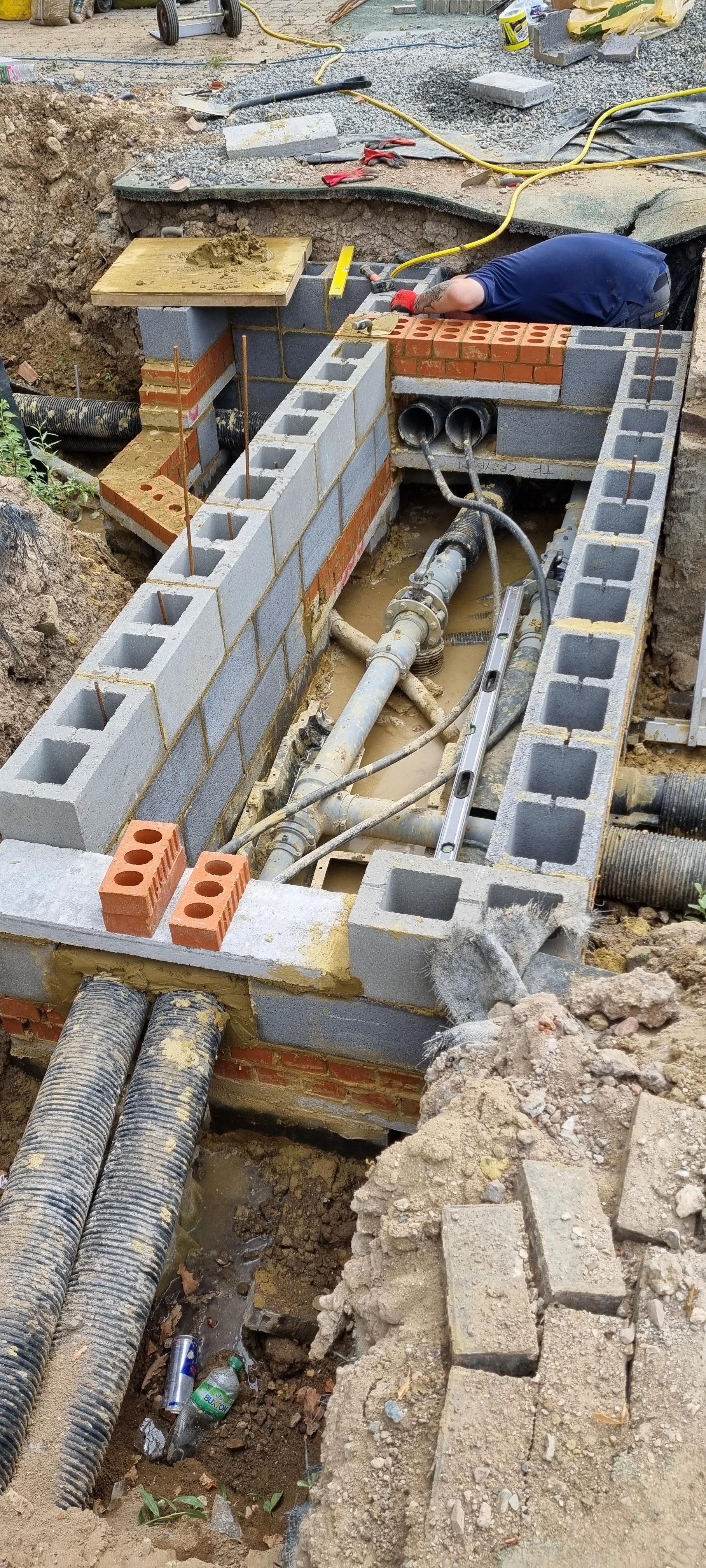 Construction site with worker laying bricks to build a wall, with pipes, cables, and tools around, and debris on the ground.