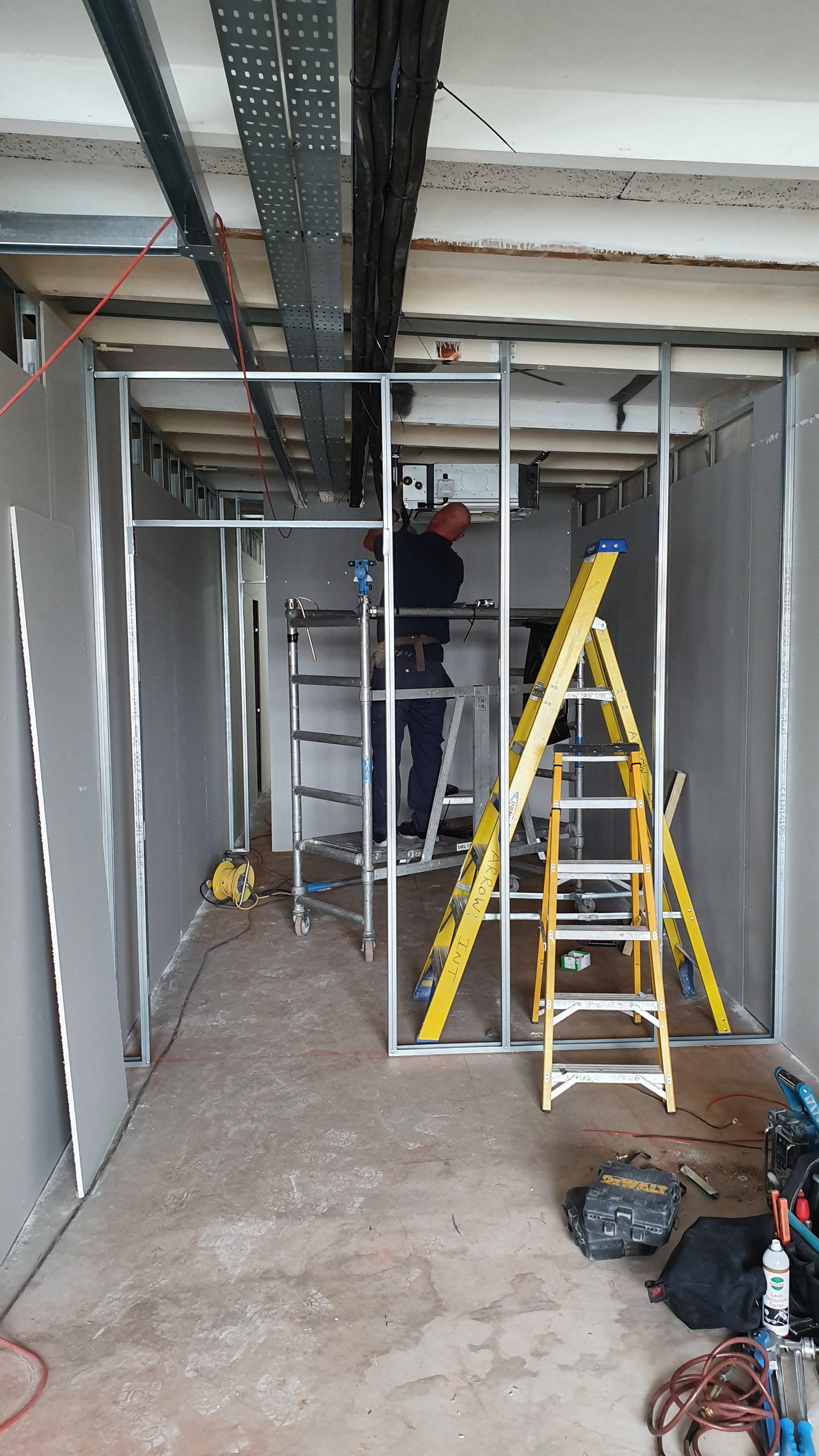 Construction worker installing electrical or HVAC system inside a framed metal room, with ladders and construction tools around.