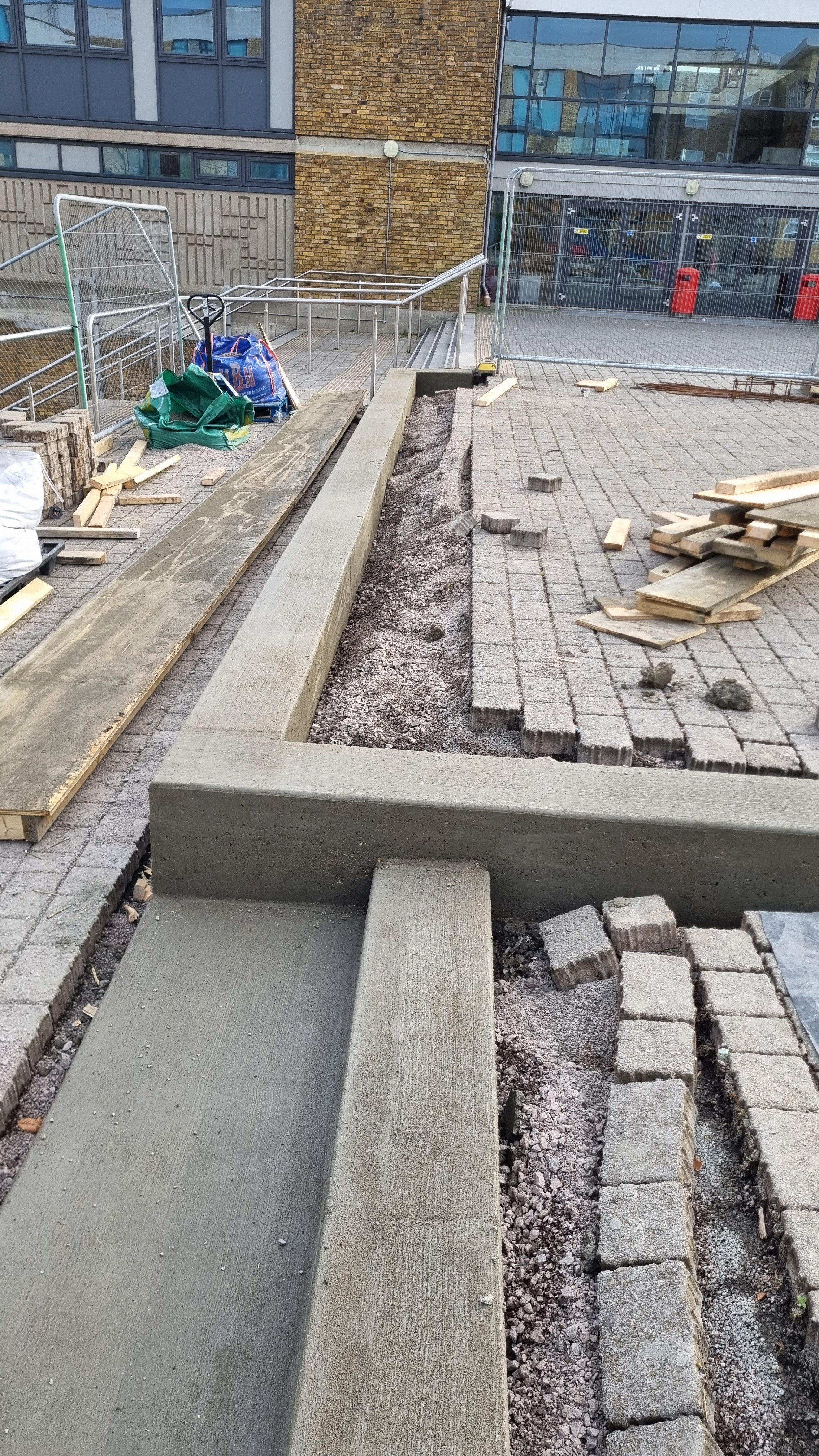 Construction workers are building a sidewalk with concrete and paving stones outside a modern building. The scene shows tools, wooden planks, and signs of ongoing work.