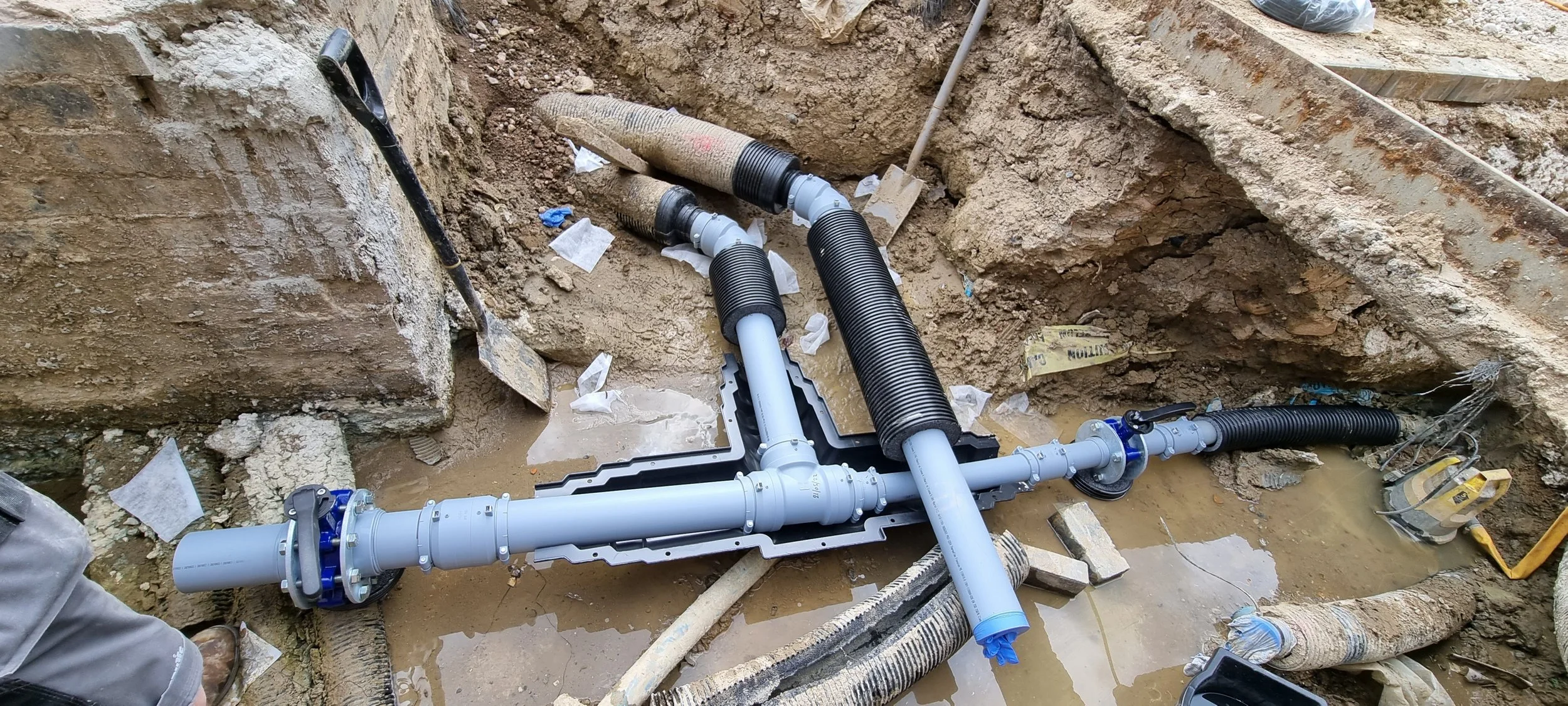 Underground plumbing pipes and fittings installed in a construction trench, with large black corrugated conduits and gray PVC pipes connected together.