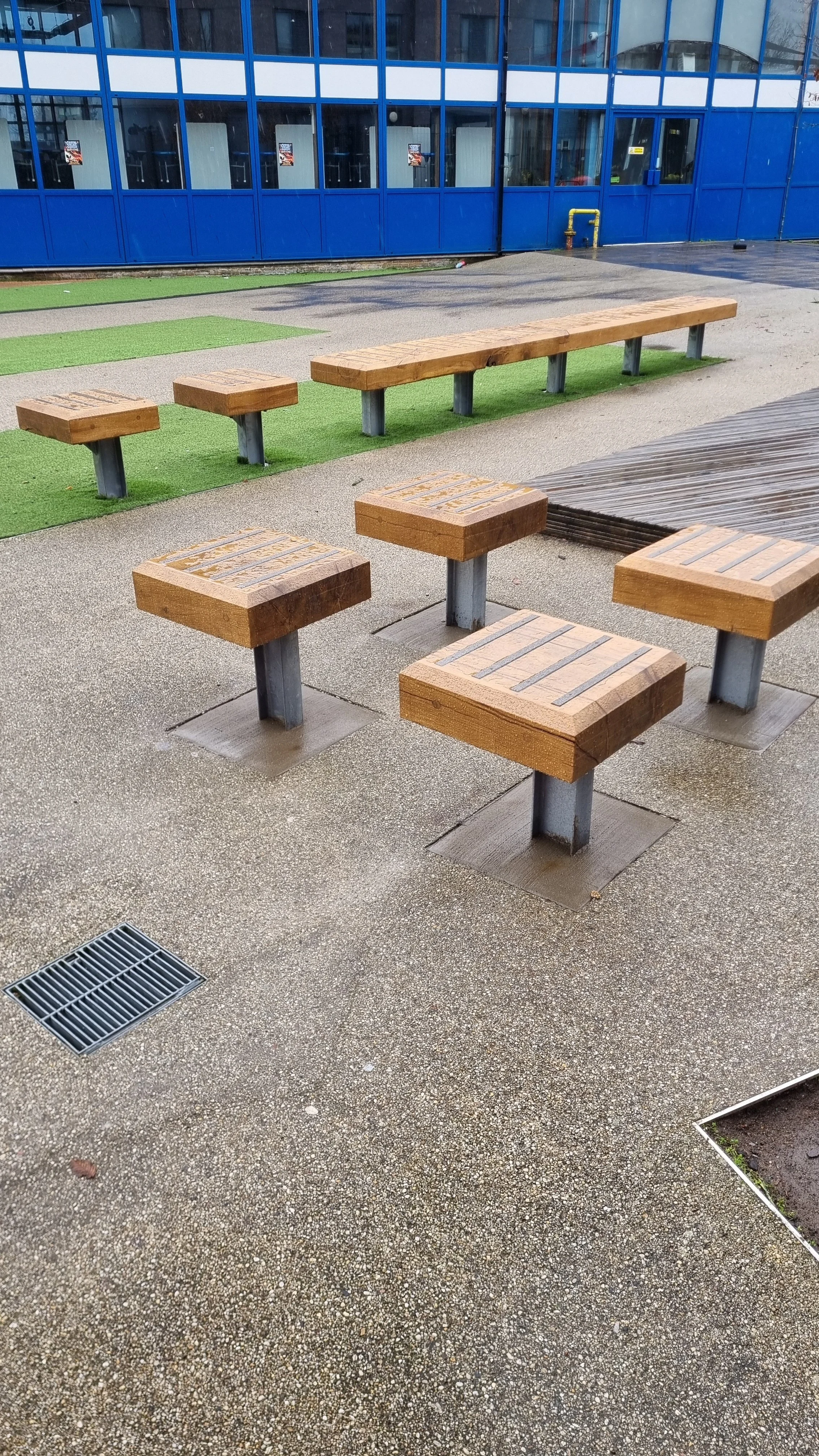 Outdoor seating area with wooden benches and square stools with metal bases on a textured concrete surface, near a blue building with large windows.