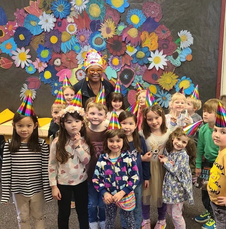 Children celebrating a birthday party, wearing colorful party hats, with a woman in the background. Some children are making peace signs, and there is a large colorful flower mural on the wall behind them.