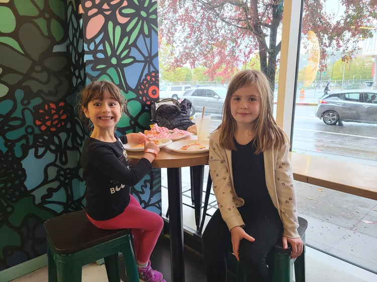 Two young girls sitting at a table inside a restaurant. One girl has a big smile, wearing a black shirt and pink pants, while the other has a neutral expression, wearing a beige hoodie and black pants. The girl with the smile has a pizza slice and a 