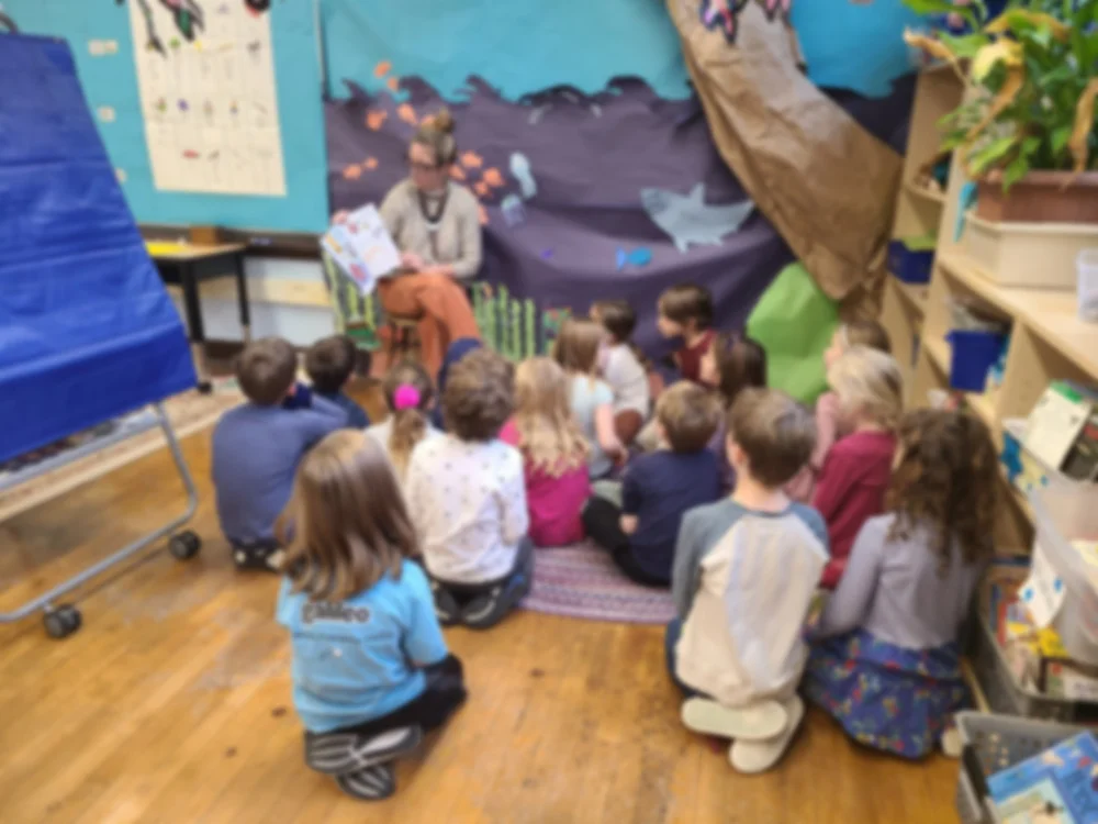 A group of children sitting on a colorful rug in a classroom, listening to a woman reading a book. The classroom has a blue and black ocean-themed mural background, with some classroom shelves and decorations visible.