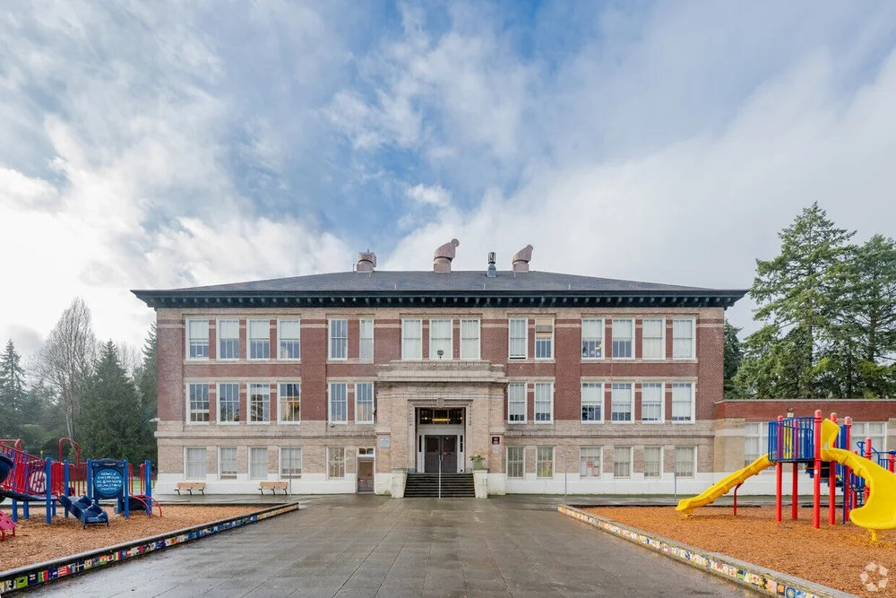 Front view of a large, three-story school building with red brick walls and white trim, flanked by colorful playground equipment on both sides, under a partly cloudy sky.