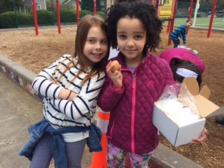 Two young girls standing together outdoors at a park, holding a donut and a box of baked goods. One girl has long brown hair and the other has curly black hair.