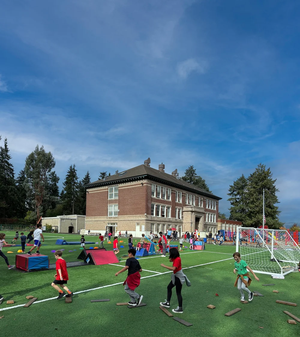 Children playing and running on a sports field during a school event with a large brick school building in the background, under a blue sky.