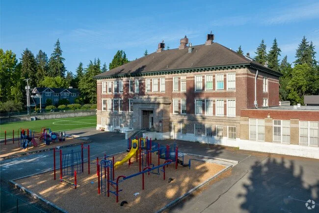 A school building with a playground in front, featuring red and blue climbing equipment and a yellow slide surrounded by a sandbox, under a clear blue sky, with trees in the background.