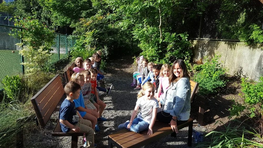 Group of children sitting on benches outdoors in a green, shaded area, some with a woman standing nearby, all smiling.