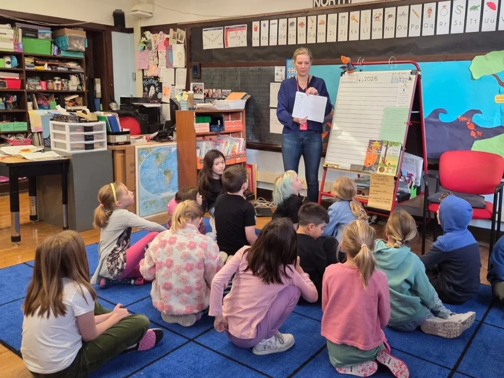 A teacher standing at a whiteboard with students seated on a blue carpet in a classroom.