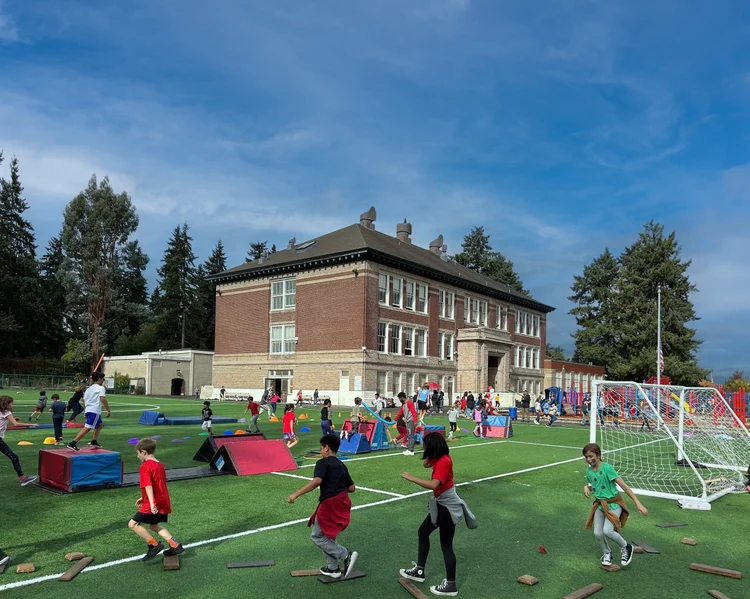Children playing on a school playground with various activities, including running, jumping, and playing with equipment, in front of a large brick school building, under a partly cloudy sky.