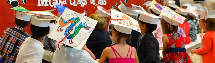 Children wearing graduation caps and colorful dresses, sitting at a long table.