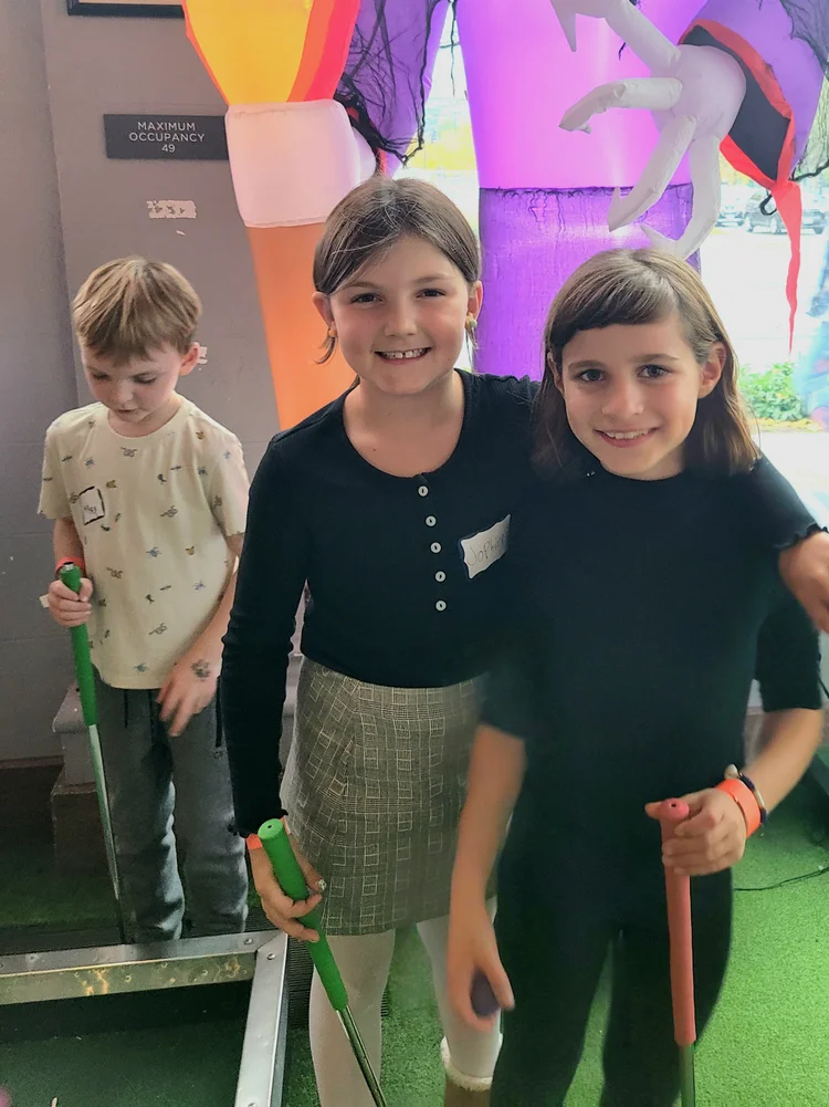 Three children in an indoor mini-golf arcade, smiling and holding mini-golf clubs. One boy with a light shirt is in the background, while two girls in dark shirts are in the foreground, standing on a green mini-golf mat. Decorations include colorful,