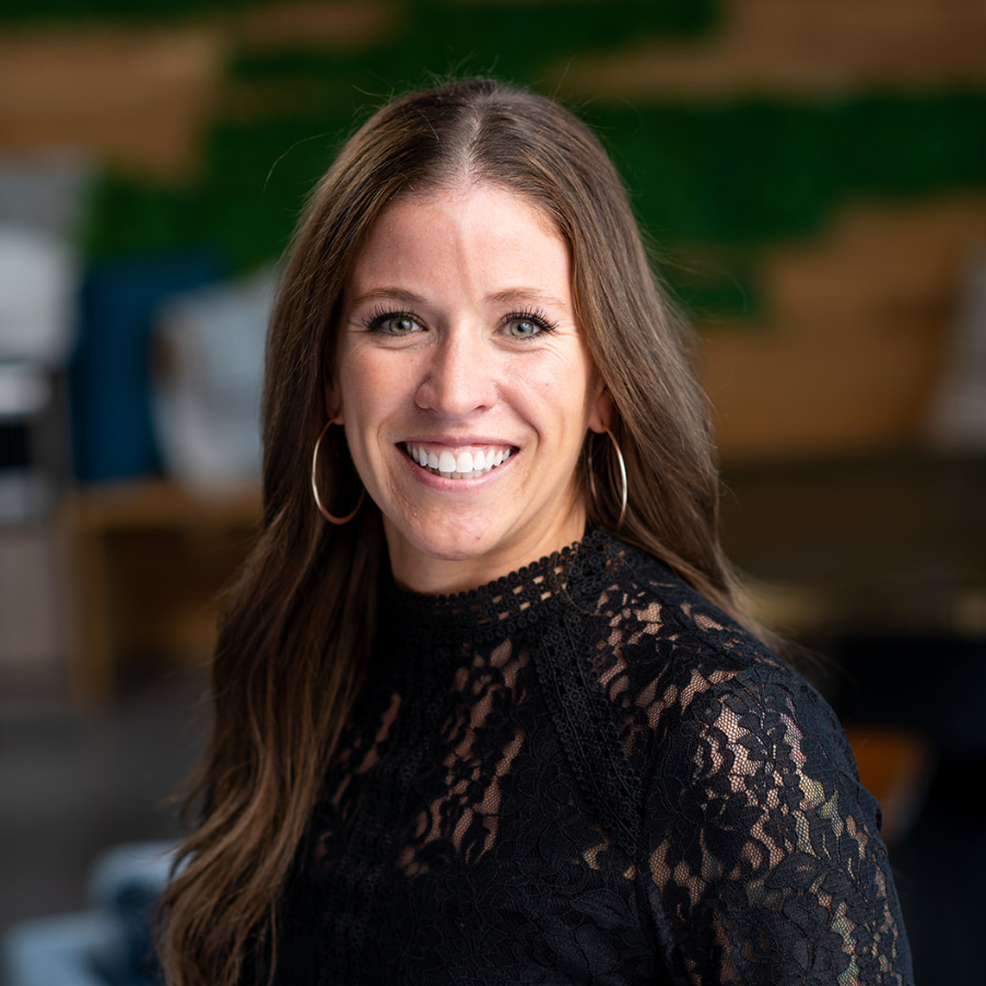 A woman with long brown hair smiling, wearing a black lace top and large hoop earrings, inside a room with wooden shelves and techno equipment in the background.