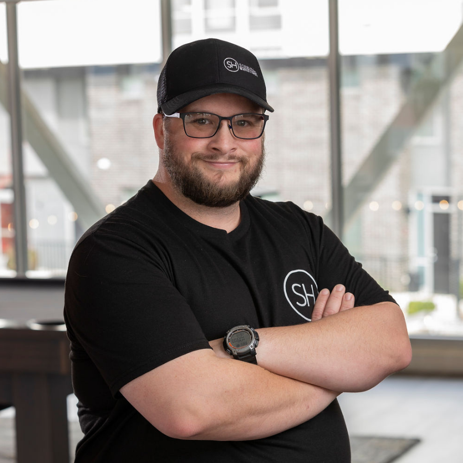 A man with glasses and a beard wearing a black cap and a black t-shirt with a logo, standing indoors with arms crossed and smiling.