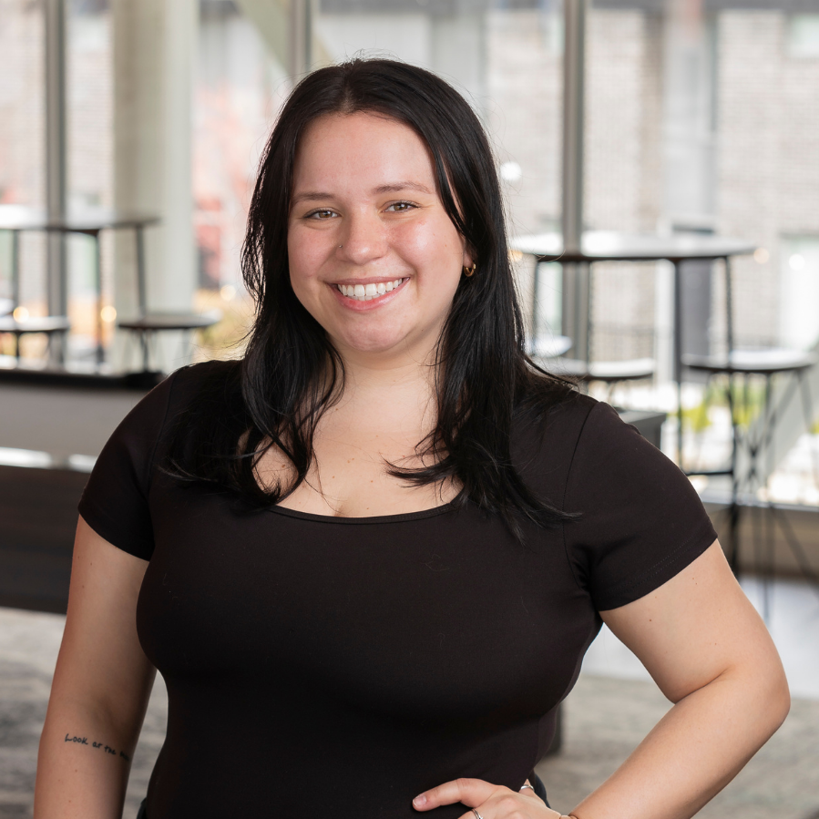 A woman with black hair smiling, wearing a black shirt, standing in a modern indoor space with large windows and a seating area in the background.