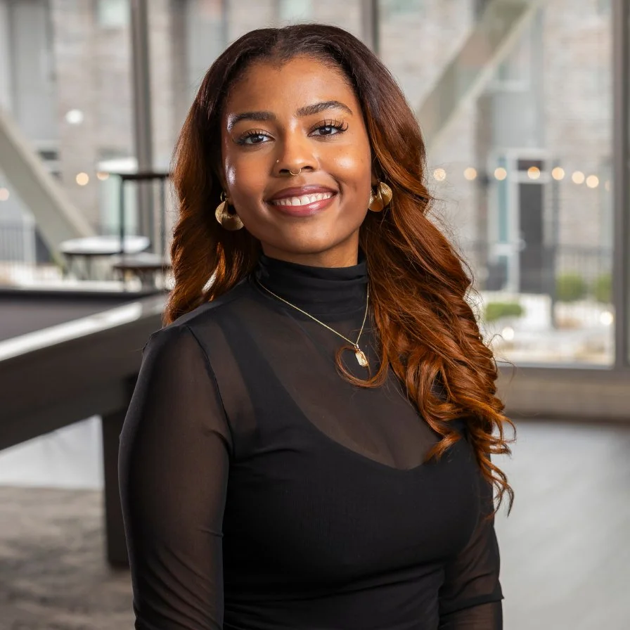 A young woman with long, wavy brown hair, wearing a black turtleneck and gold jewelry, smiling indoors with large windows and cityscape background.