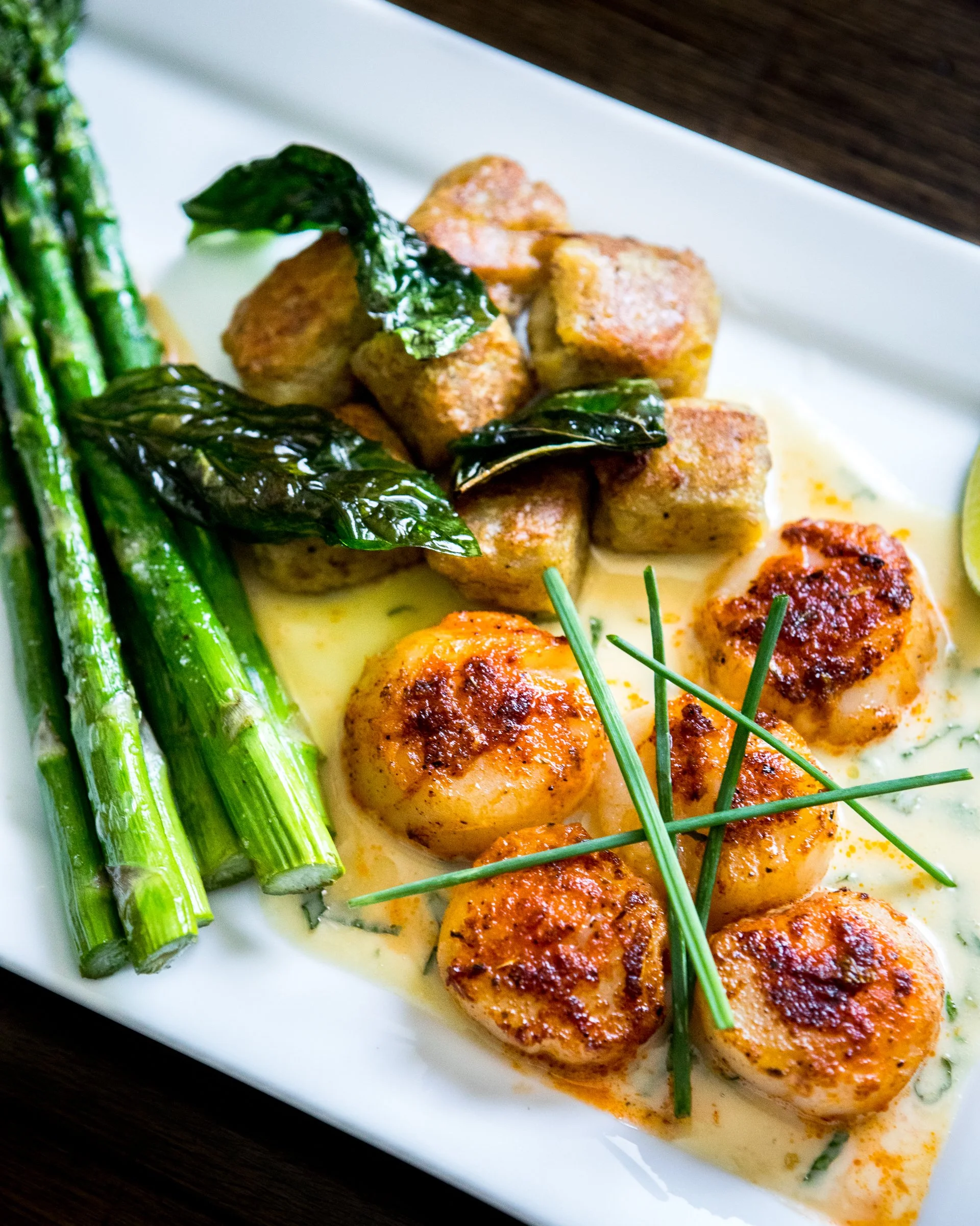 A white plate with sautéed scallops in creamy sauce, grilled asparagus, fried hash brown cubes topped with basil leaves, and lime wedge.
