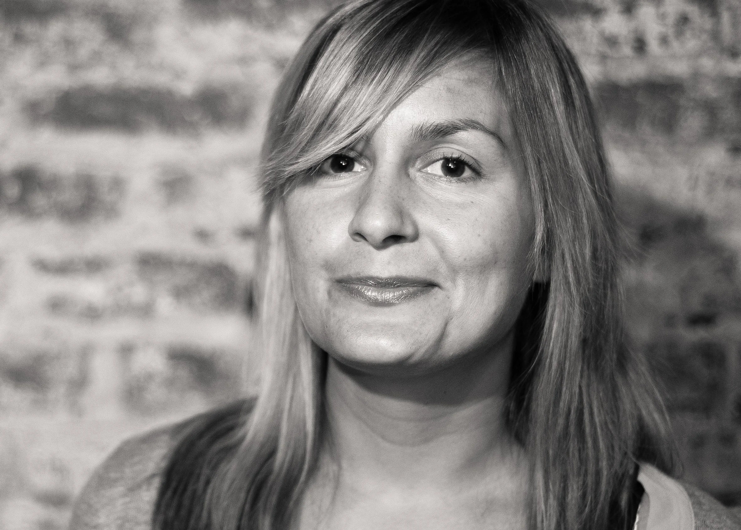 A black and white portrait of a woman with long, light-colored hair, standing in front of a brick wall.