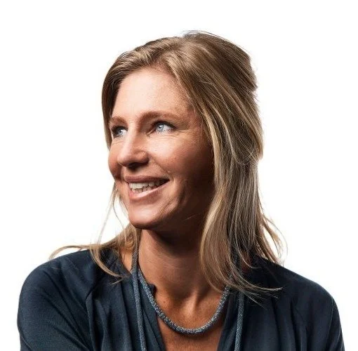 A woman with blonde hair smiling and looking to her right, wearing a dark top and a necklace.