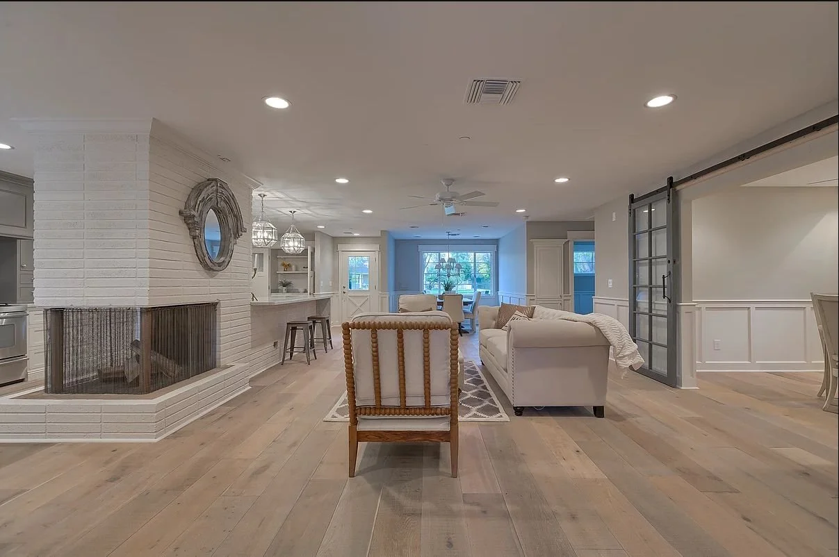 Open concept living room and kitchen with a modern design, featuring light-colored hardwood floors, a central brick fireplace, a cozy seating area with sofas, a kitchen counter with bar stools, and recessed ceiling lights. A sliding barn door is visible on the right.