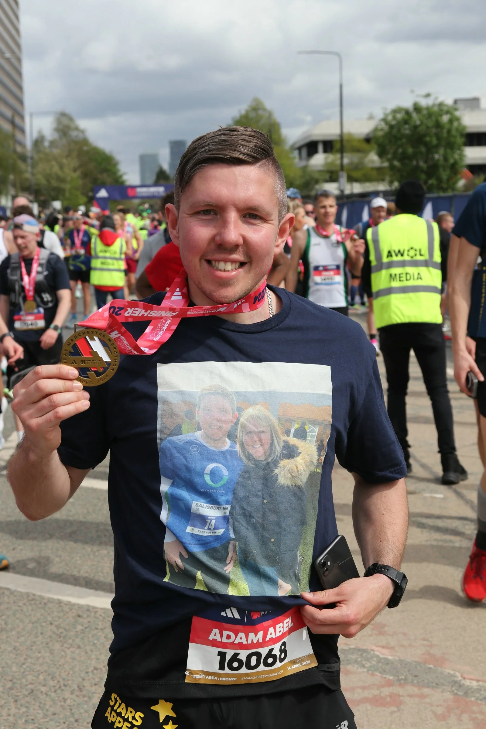 Adam Abe holding his medal at Manchester Marathon in a crowd of runners and event staff.
